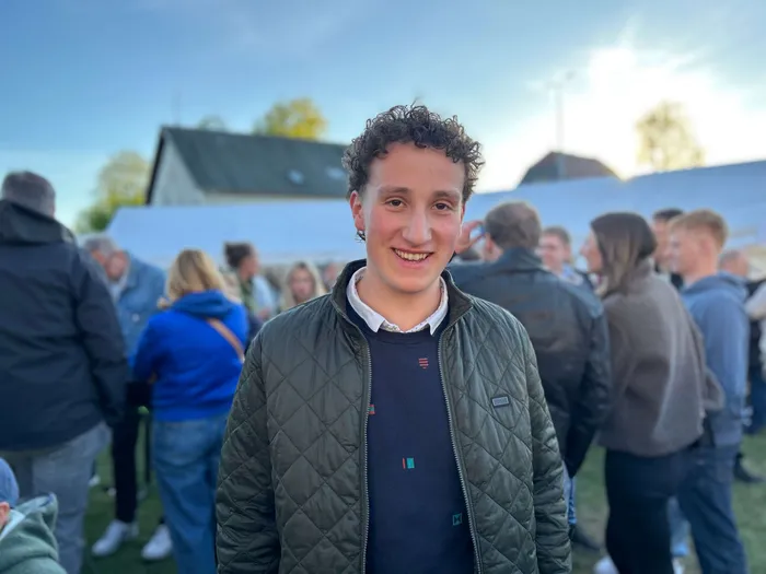 Ein junger Mann mit lockigem Haar lächelt in die Kamera, im Hintergrund Menschen auf einem Dorfplatz während eines Maibaumfestes.