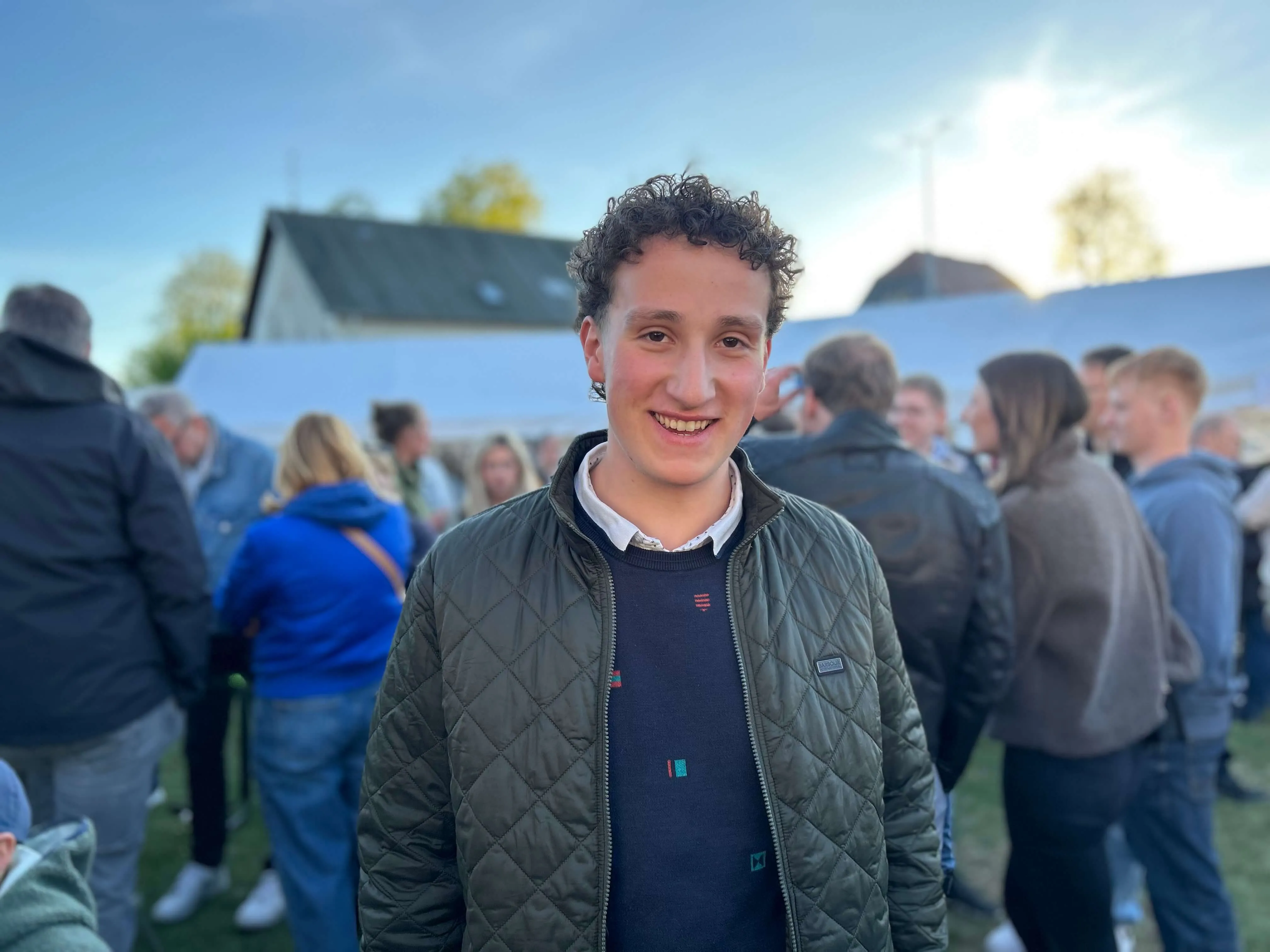 Ein junger Mann mit lockigem Haar lächelt in die Kamera, im Hintergrund Menschen auf einem Dorfplatz während eines Maibaumfestes.