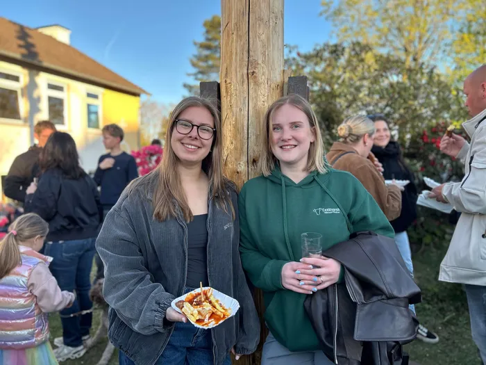 Zwei junge Frauen lächeln in die Kamera, während sie auf einem Frühlingsfest Pommes aus einer Pappschale essen. Im Hintergrund sind weitere Menschen und ein Gebäude zu sehen.