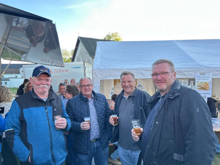 Drei Männer mittleren Alters stehen auf einem Festgelände zusammen und halten Biergläser in der Hand. Im Hintergrund ein Festzelt und ein Banner mit der Aufschrift „Gox & Söhne“.