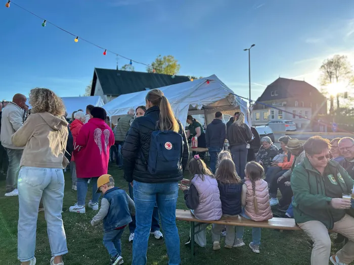 Menschengruppen stehen und sitzen auf einer Wiese vor einem großen Zelt und einem Gebäude mit Satteldach. Lichterketten sind über dem Gelände gespannt.