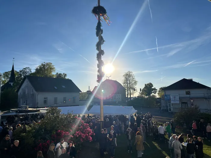 Ein hoher, geschmückter Maibaum steht im Zentrum eines Festplatzes, umgeben von zahlreichen Menschen und Gebäuden mit traditioneller Architektur.