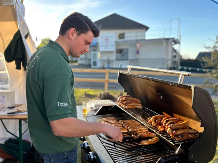 Ein Mann steht an einem Grill und brät Würstchen. Im Hintergrund ein Gebäude und eine Holzbarriere.