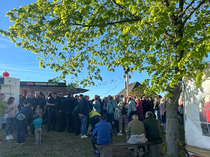 Menschen stehen und sitzen vor einem roten Imbissstand mit der Aufschrift „Linnepe Liere“. Im Hintergrund ist ein Maibaum mit bunten Bändern zu sehen.