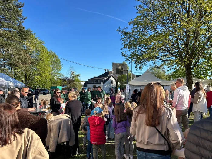 Menschengruppen stehen und unterhalten sich vor einem traditionellen Fachwerkhaus und mehreren weißen Zelten auf einer grünen Wiese. Im Hintergrund blauer Himmel und Bäume.