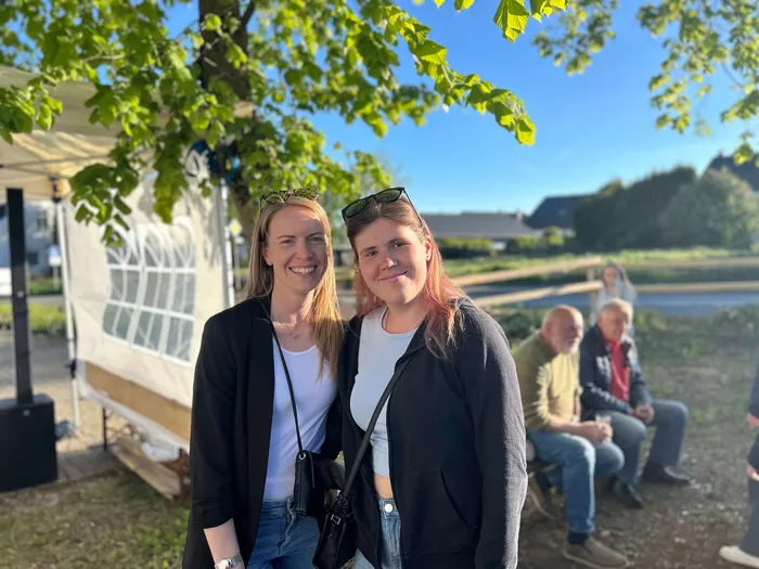 Zwei junge Frauen lächeln in die Kamera, vor ihnen ein Festzelt und im Hintergrund eine grüne Landschaft mit Häusern.