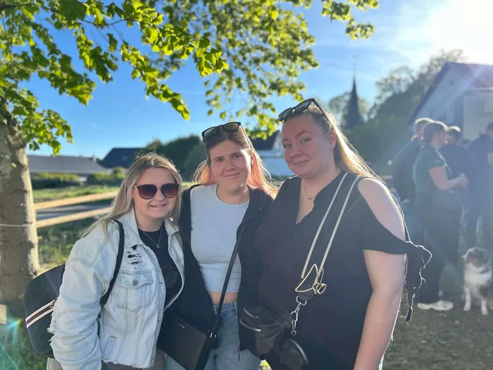 Drei junge Frauen lächeln in die Kamera, im Hintergrund Menschen und ein Gebäude mit spitzem Dach. Das Bild wurde offenbar bei einem Maibaumfest aufgenommen.