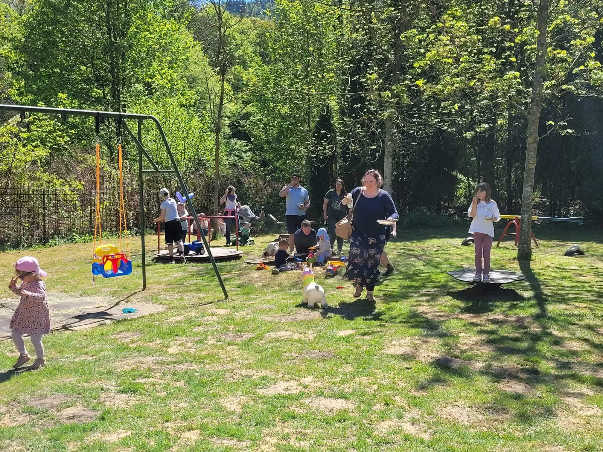 Menschen unterschiedlichen Alters feiern im Freien auf einer Wiese, einige sitzen an Tischen, andere spielen oder stehen um eine Schaukel und eine Rutsche.