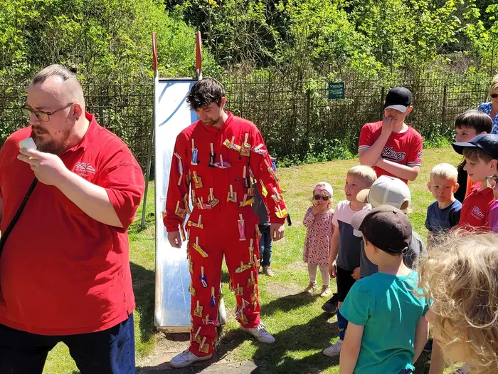 Ein Mann in roter Kleidung schneidet ein Stück Kuchen, während eine Gruppe von Kindern und Erwachsenen zusieht. Im Hintergrund steht ein weißes Schild und Bäume.