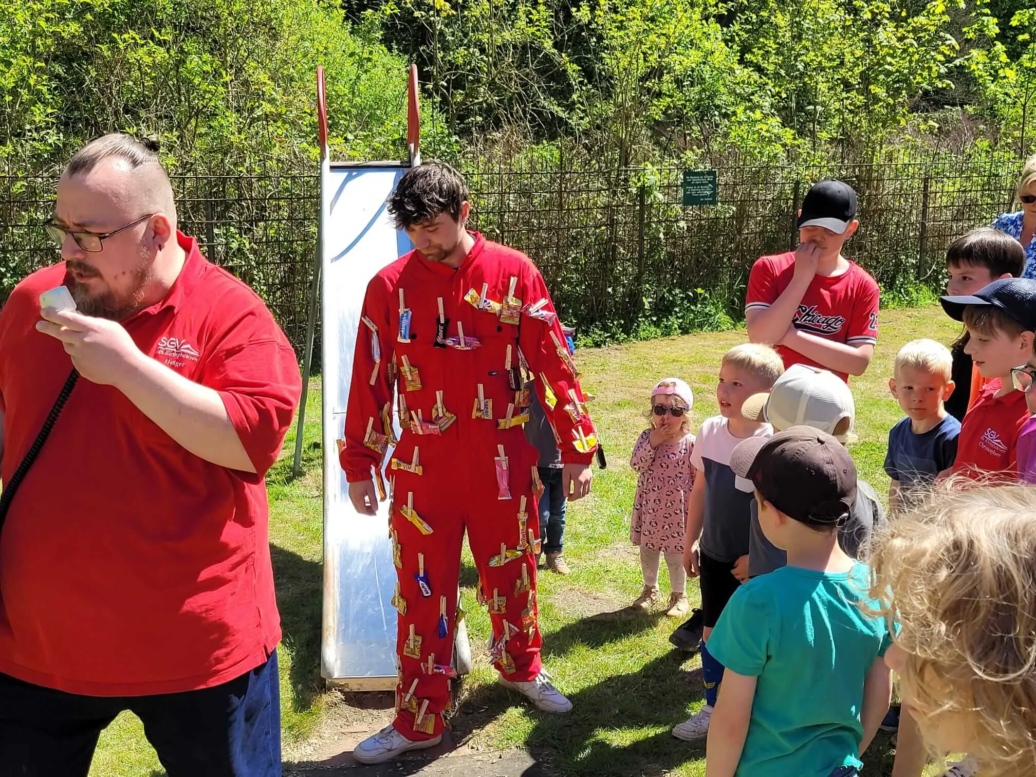 Ein Mann in roter Kleidung schneidet ein Stück Kuchen, während eine Gruppe von Kindern und Erwachsenen zusieht. Im Hintergrund steht ein weißes Schild und Bäume.