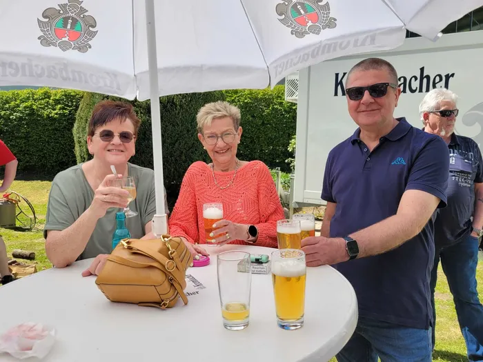 Draußen sitzen Menschen an einem Tisch unter Sonnenschirmen und trinken Bier. Im Hintergrund weitere Besucher und ein Schild mit der Aufschrift „Kocher“.