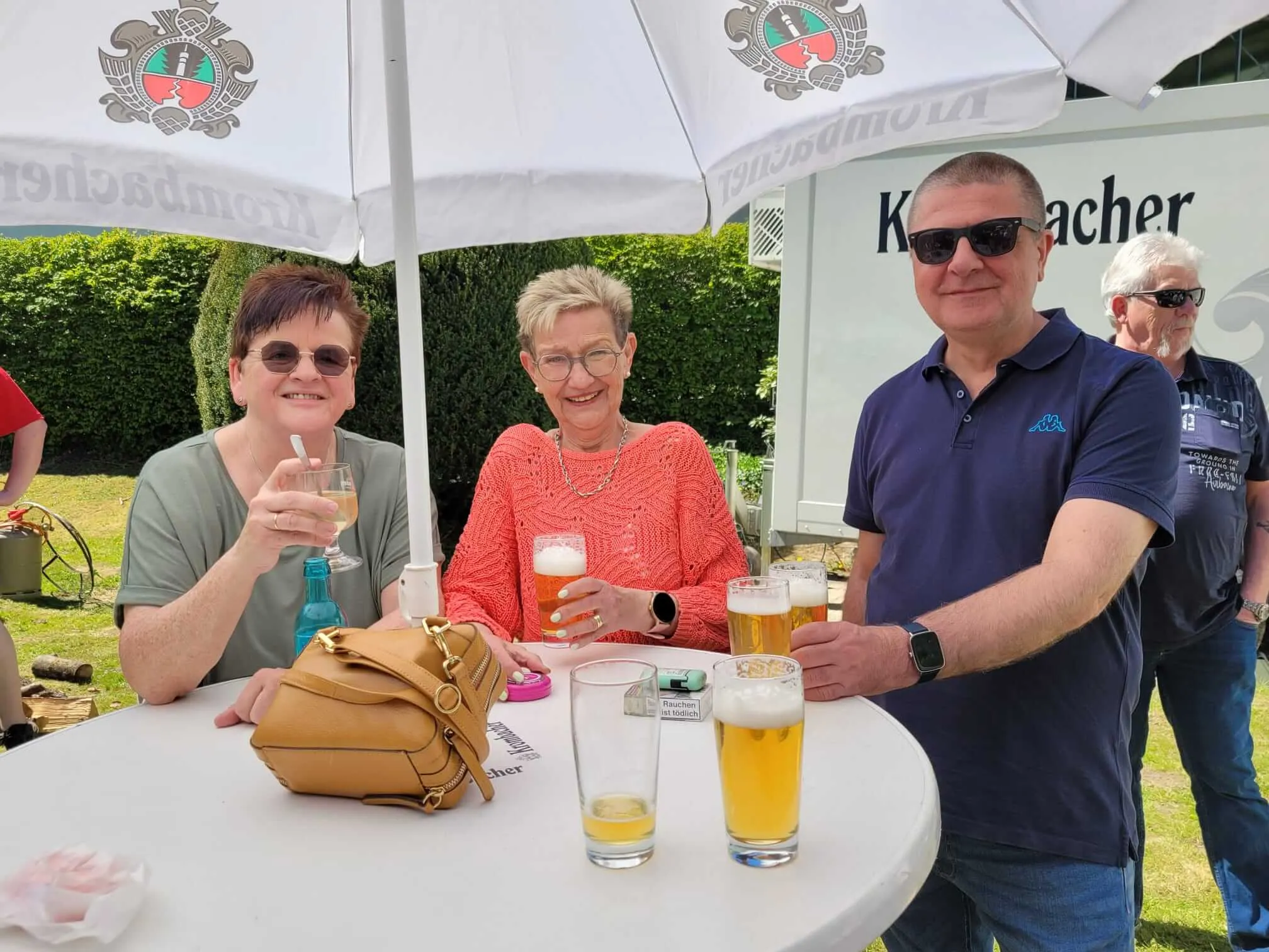 Draußen sitzen Menschen an einem Tisch unter Sonnenschirmen und trinken Bier. Im Hintergrund weitere Besucher und ein Schild mit der Aufschrift „Kocher“.