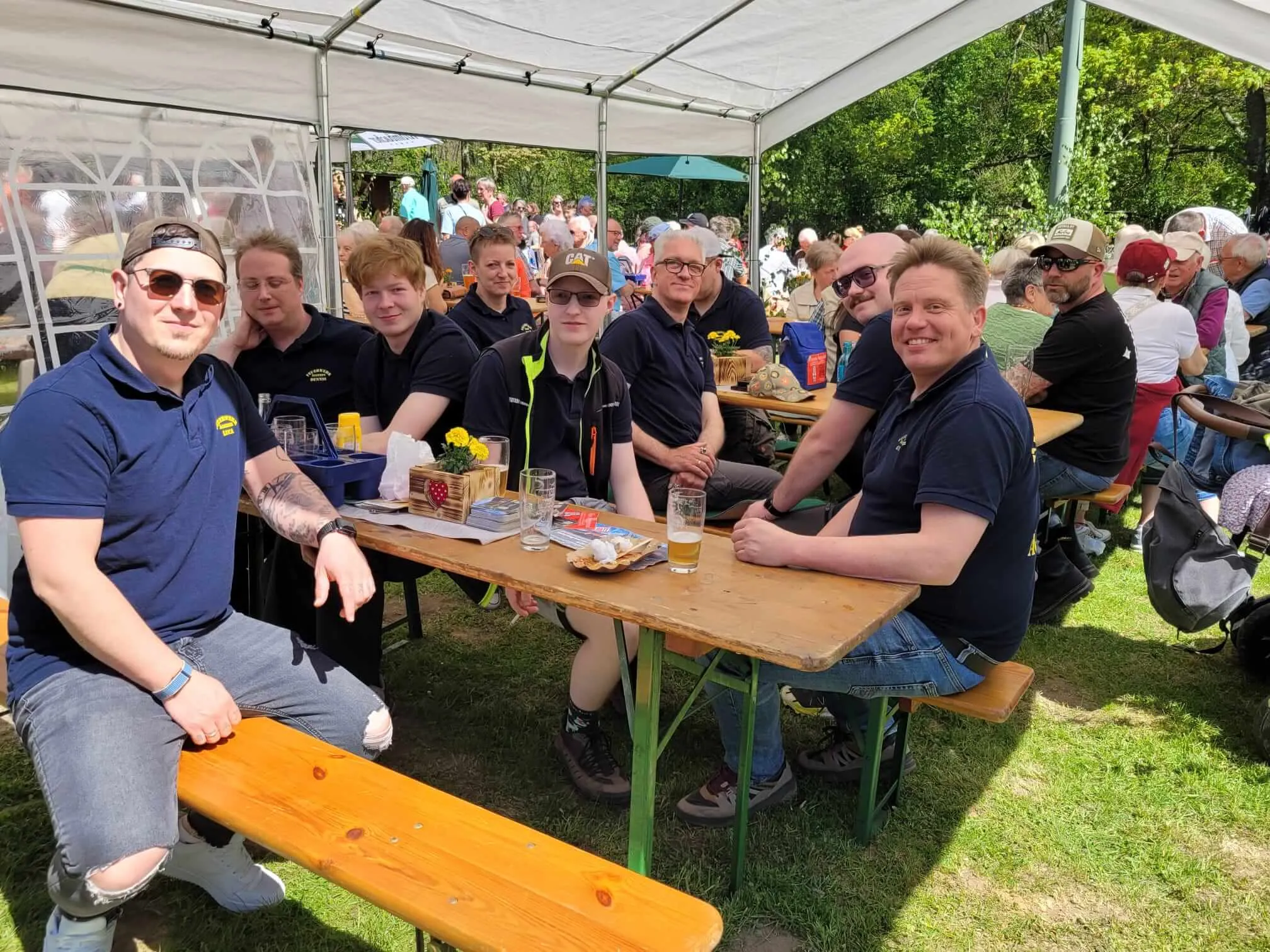 Eine Gruppe von Menschen sitzt an einem Biergartentisch unter einem Zelt und genießt Getränke und Gespräche bei einer Maifeier.