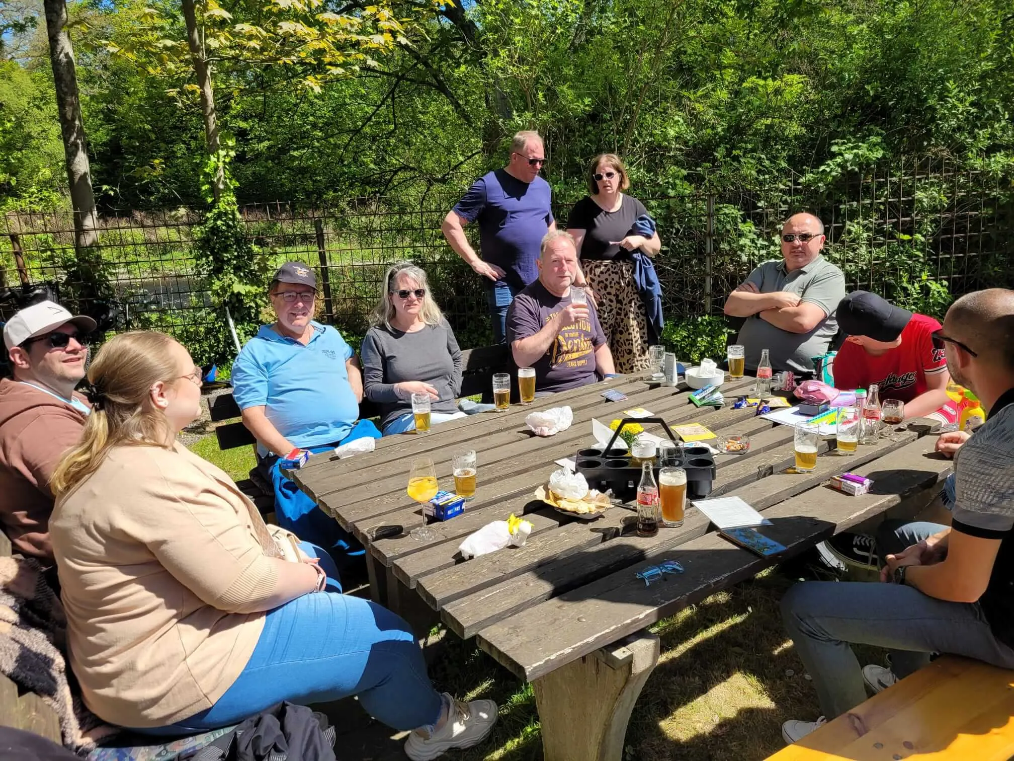 Eine Gruppe von Menschen sitzt und steht um einen langen Picknicktisch im Freien, Getränke und Snacks darauf. Im Hintergrund sind Bäume und ein Zaun zu sehen.