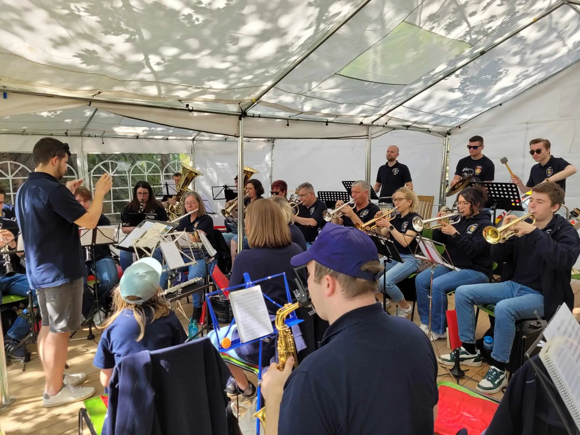 Blasorchester spielt unter einem Zelt im Freien, viele Musiker und Zuschauer sind zu sehen.