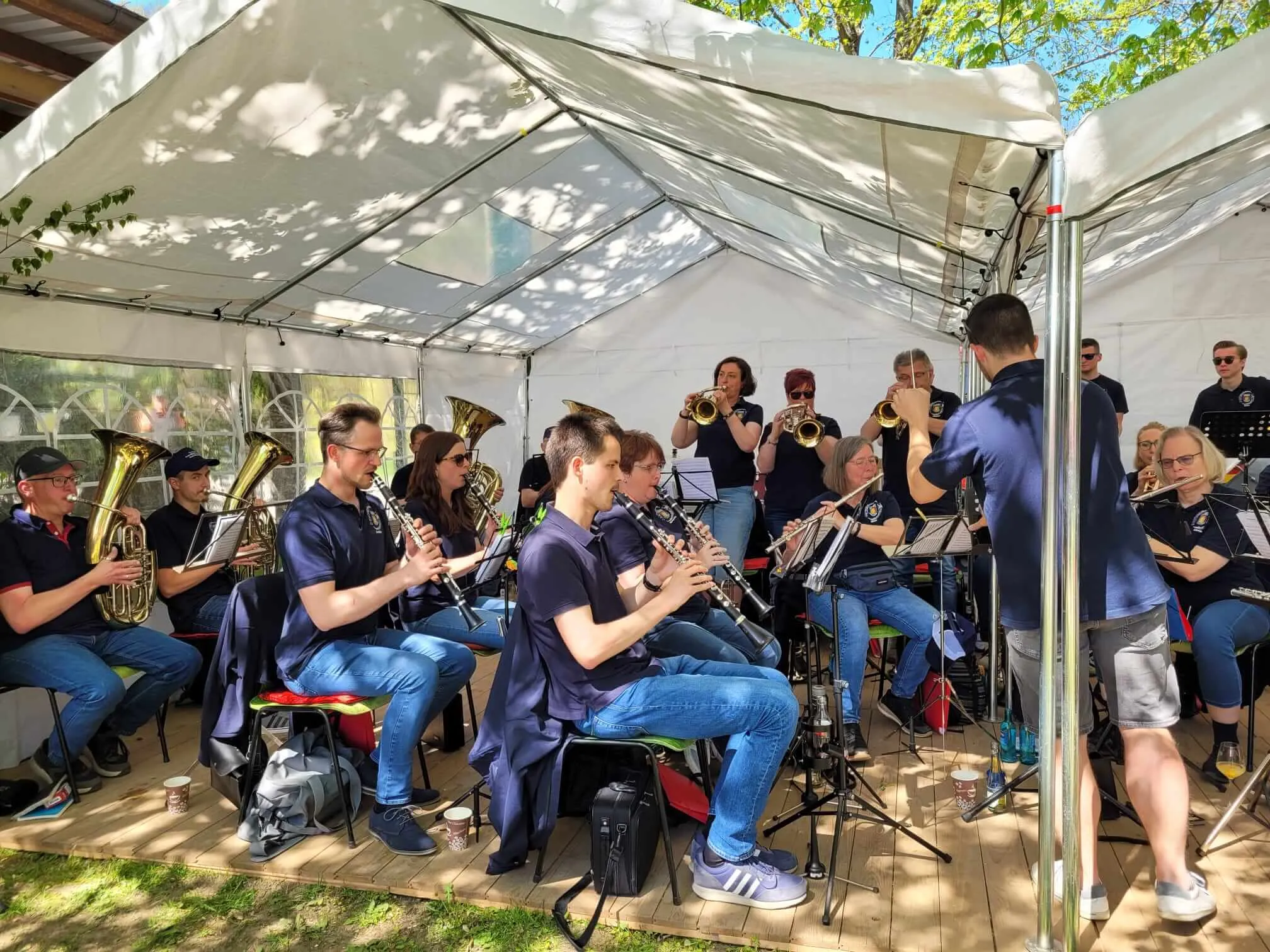 Blasorchester spielt unter einem Zelt im Freien. Mehrere Musiker spielen verschiedene Instrumente wie Posaune, Klarinette und Trompete.