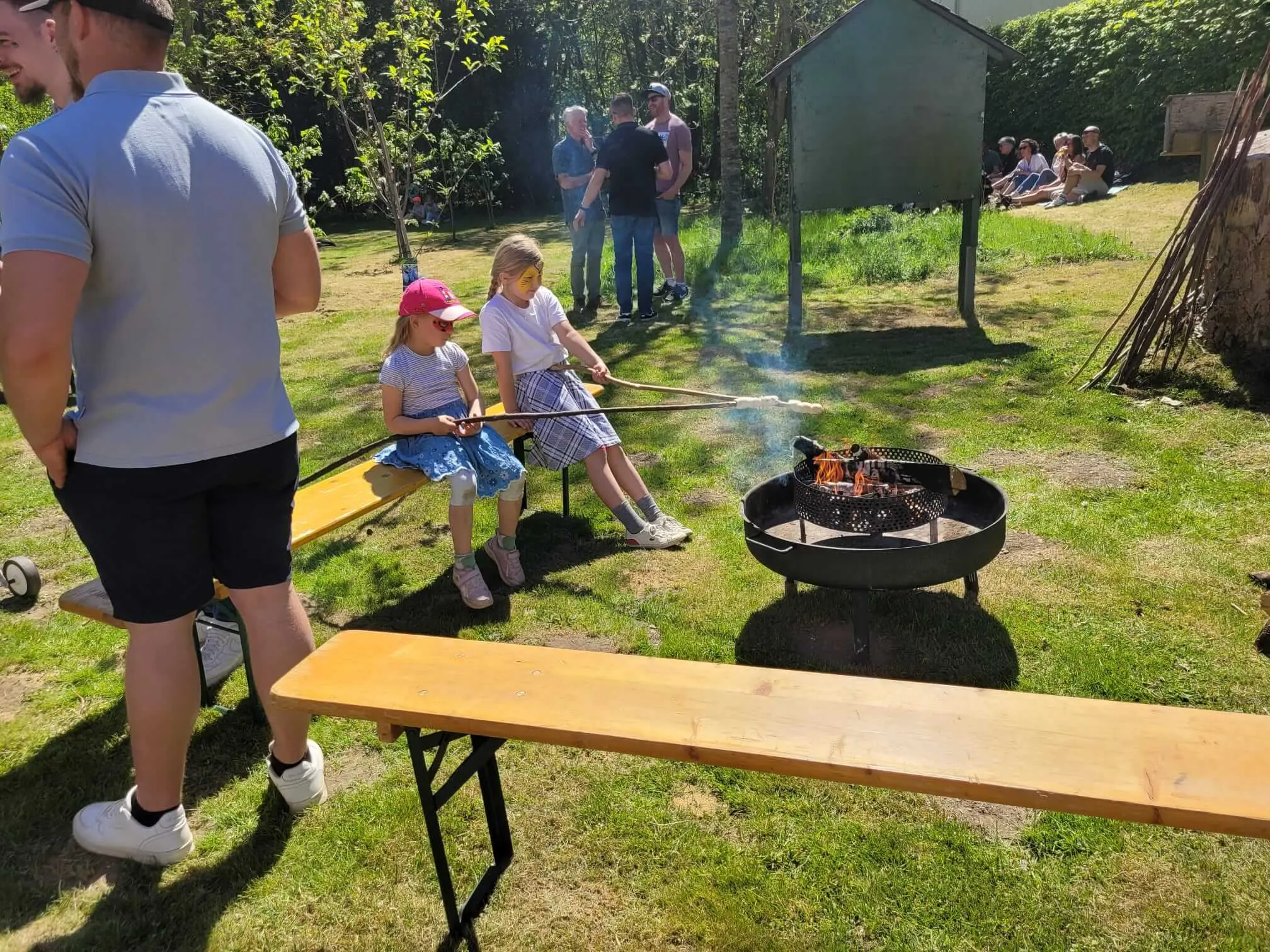 Zwei Kinder stecken Marshmallows über einem Lagerfeuer auf einer Wiese, während im Hintergrund Menschen stehen und sitzen.