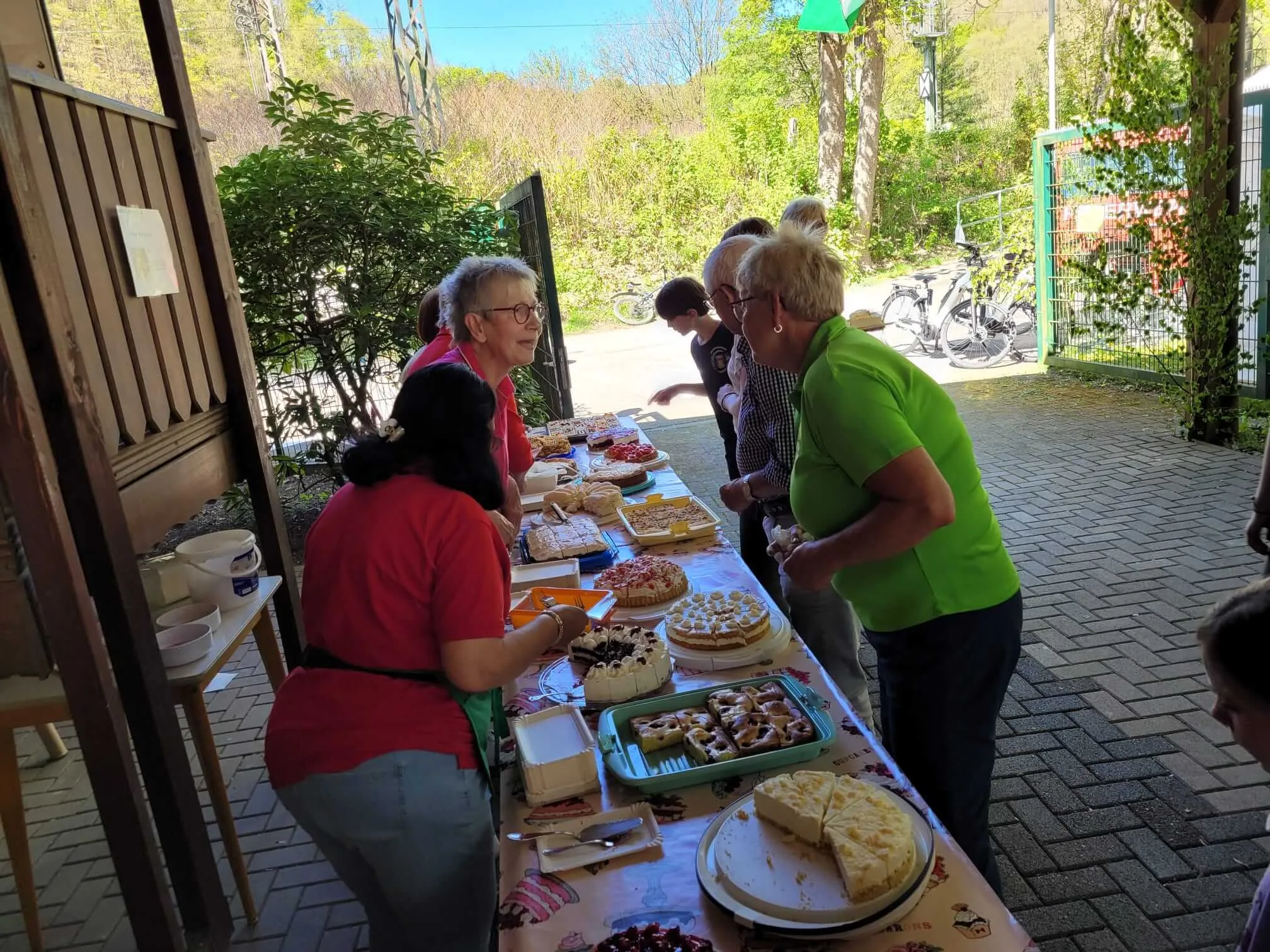 Frauen bedienen einen Tisch mit Kuchen und Gebäck im Freien, während andere Besucher im Hintergrund stehen.