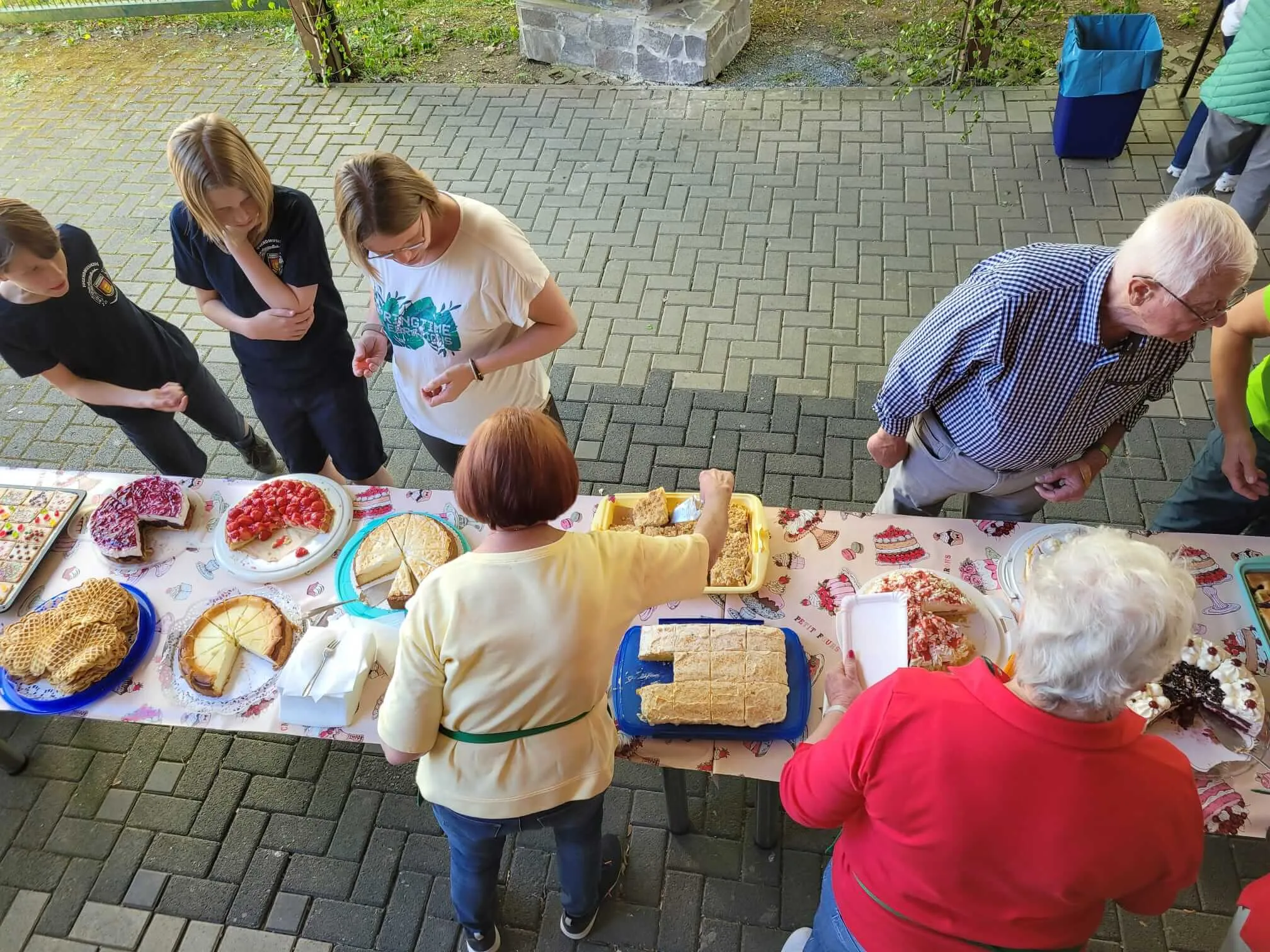 Menschen stehen um gefüllte Tische mit Kuchen und Gebäck im Freien. Im Hintergrund ein Mann mit blauem Hemd und eine Frau mit rotem Oberteil.