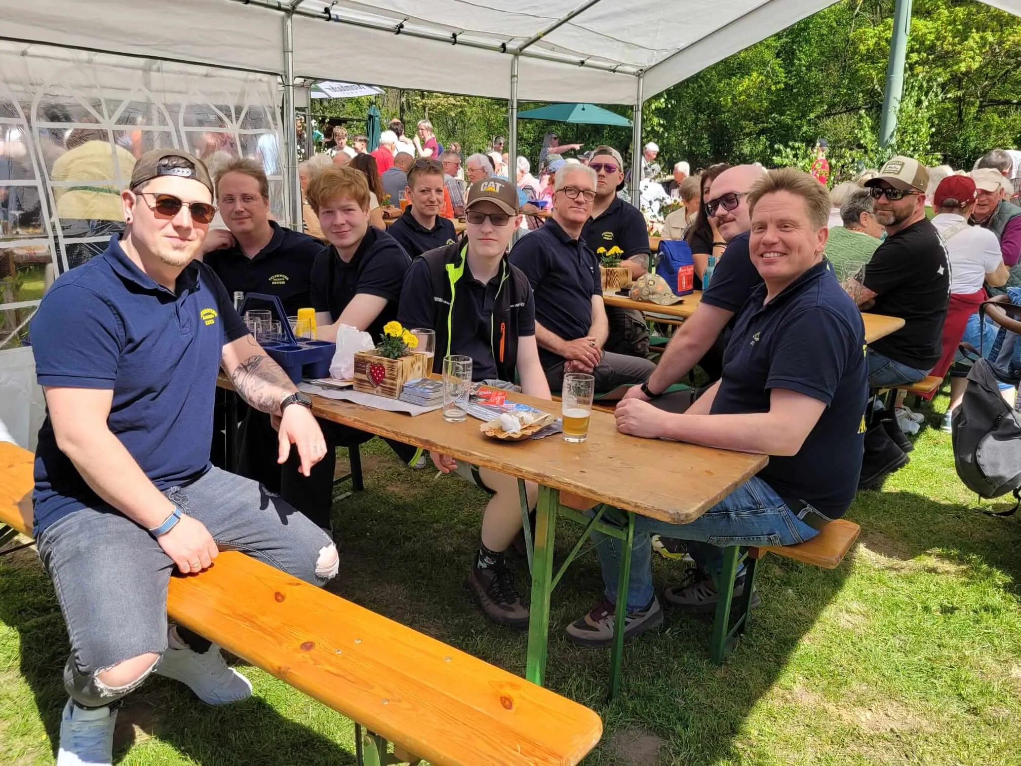 Eine Gruppe von Menschen sitzt an Biertischbänken im Freien, einige halten Getränke. Im Hintergrund sind weitere Besucher und Zelte auf einer Maifeier zu sehen.