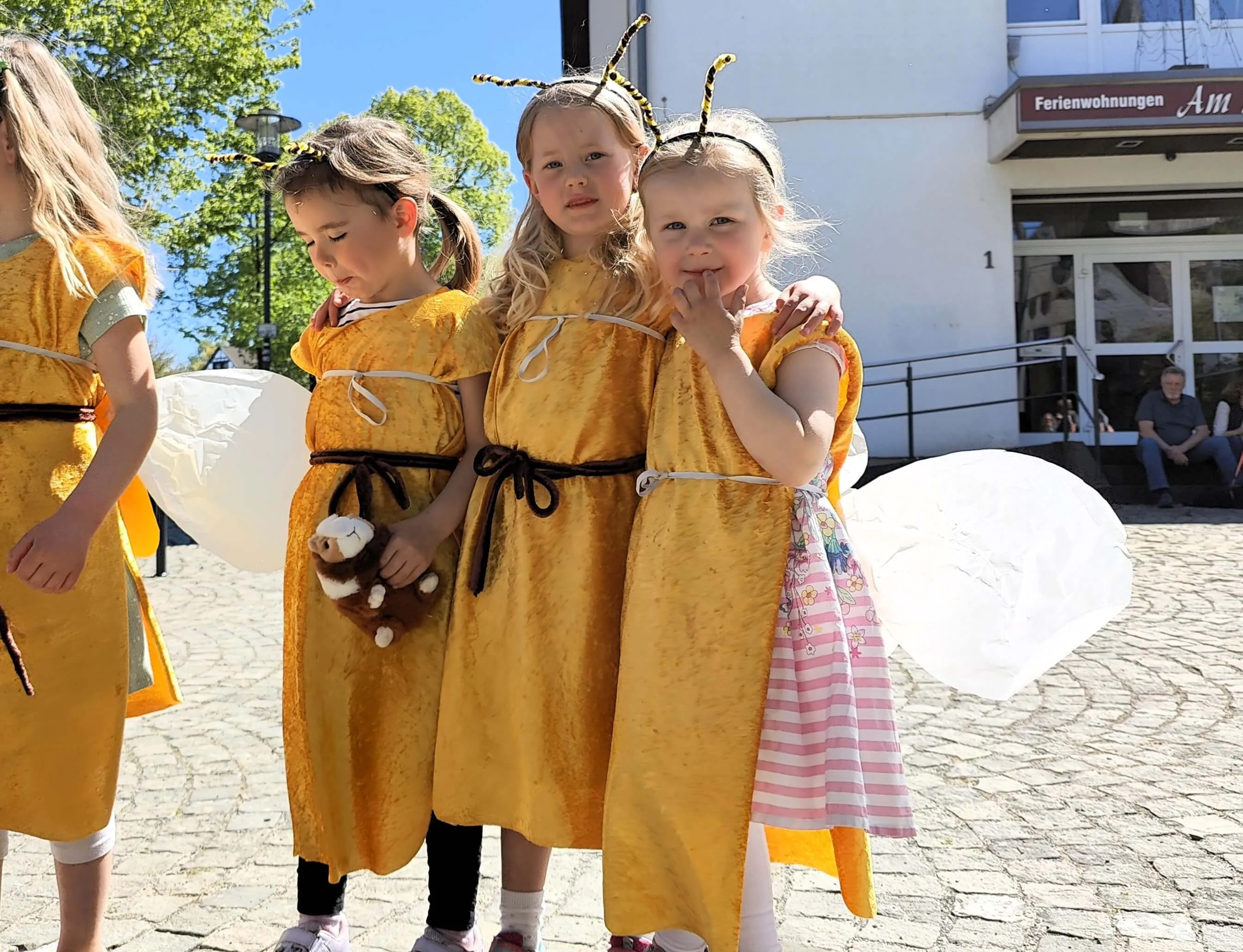 Vier Kinder in Bienenkostümen mit Flügeln stehen auf einem Platz vor einem Gebäude. Sie tragen gelbe Kleider und Kopfbedeckungen mit Fühlern.
