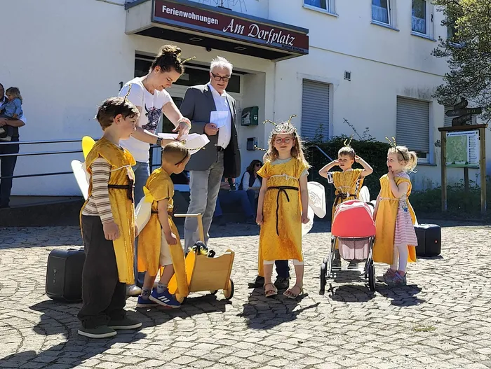 Eine Gruppe von Kindern in Bienenkostümen steht vor einem Mann im Anzug, während eine Frau einem Kind eine Flügel befestigt. Im Hintergrund ein Gebäude mit dem Schild „Ferienwohnungen Am Dorfplatz“.