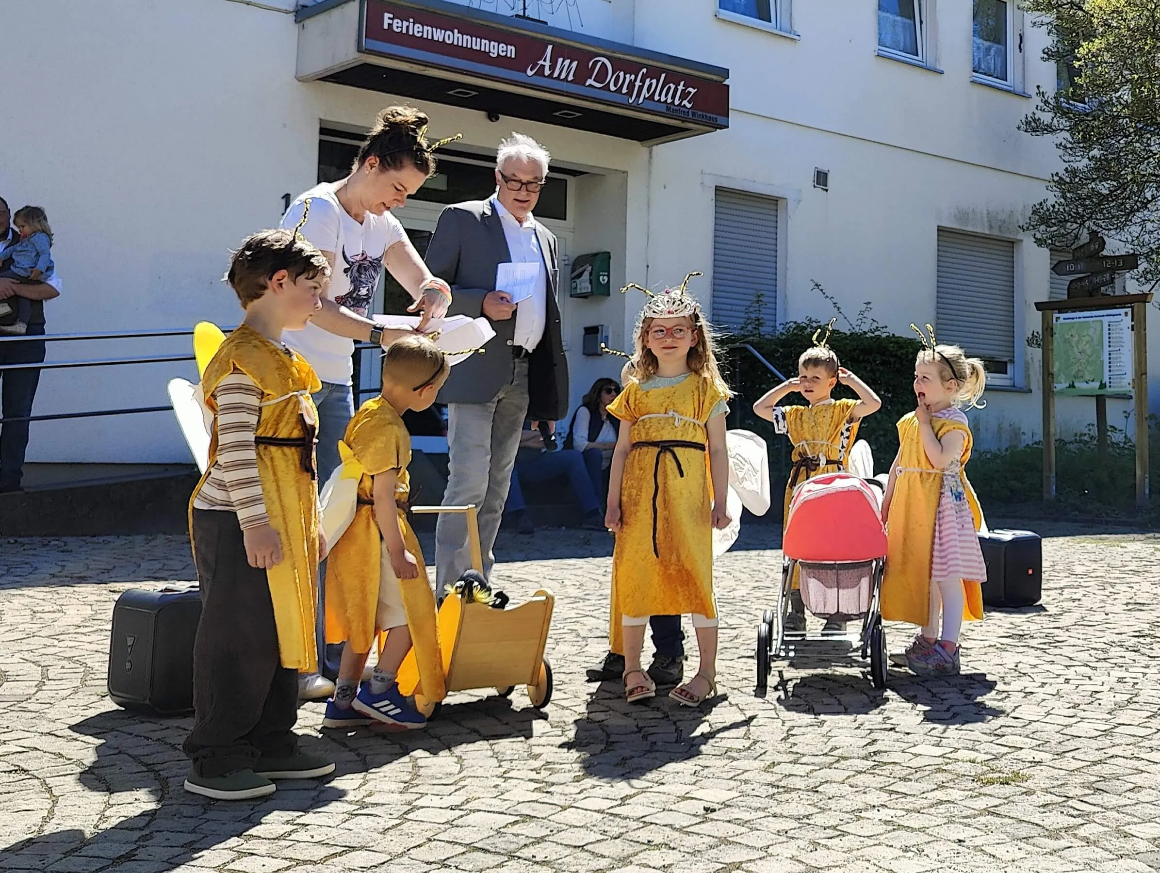 Eine Gruppe von Kindern in Bienenkostümen steht vor einem Mann im Anzug, während eine Frau einem Kind eine Flügel befestigt. Im Hintergrund ein Gebäude mit dem Schild „Ferienwohnungen Am Dorfplatz“.