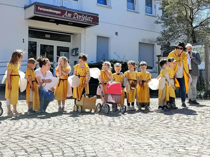 Eine Gruppe Kinder in gelben Gewändern steht auf einem gepflasterten Platz vor einem hellen Gebäude mit dem Schild „Ferienwohnungen Am Dorfplatz“. Eine Frau und ein Mann begleiten die Kinder.