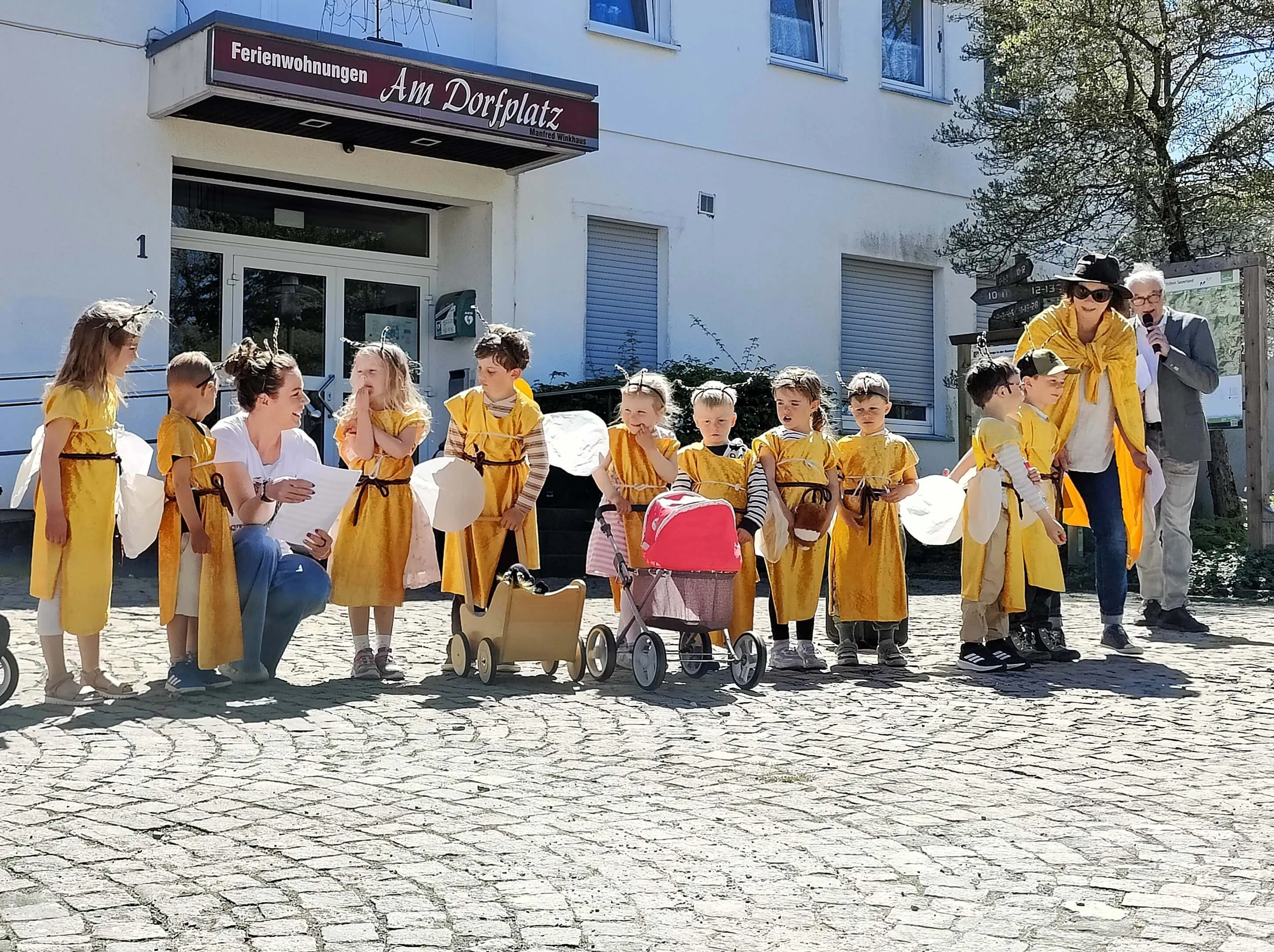 Eine Gruppe Kinder in gelben Gewändern steht auf einem gepflasterten Platz vor einem hellen Gebäude mit dem Schild „Ferienwohnungen Am Dorfplatz“. Eine Frau und ein Mann begleiten die Kinder.
