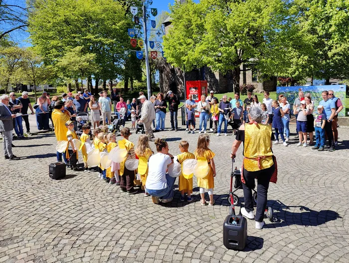 Kinder in gelben Kleidern mit weißen Flügeln tanzen im Kreis um eine Frau, umgeben von Zuschauern auf dem Dorfplatz Wiblingwerde.