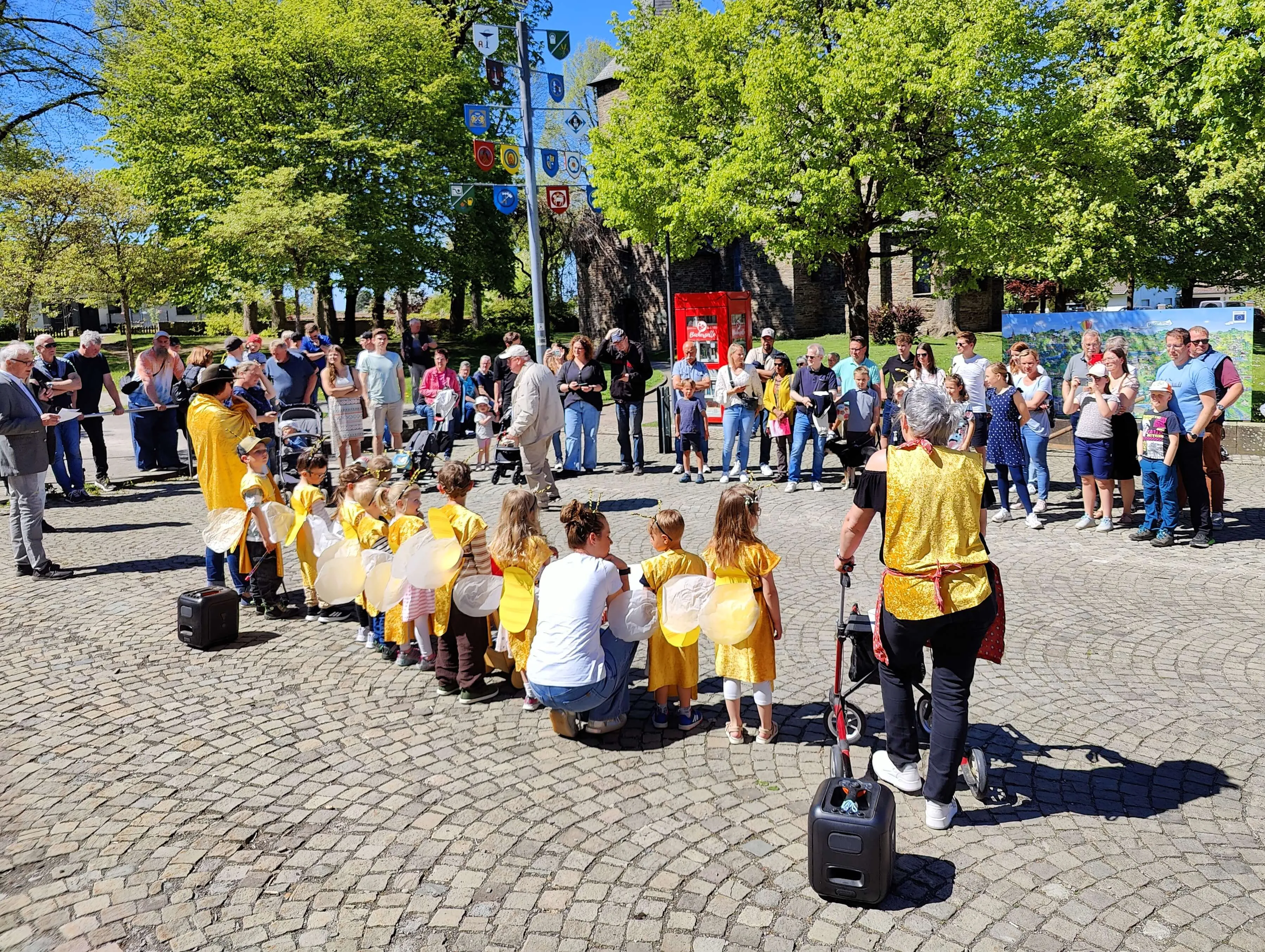 Kinder in gelben Kleidern mit weißen Flügeln tanzen im Kreis um eine Frau, umgeben von Zuschauern auf dem Dorfplatz Wiblingwerde.