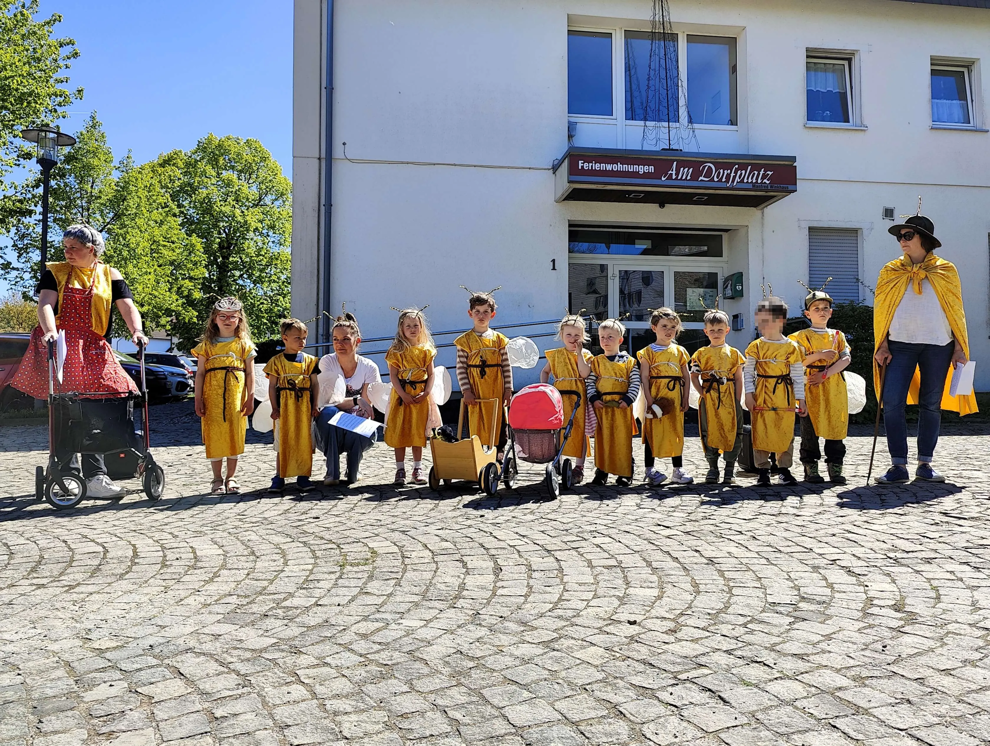 Eine Gruppe von Kindern in gelben Kostümen mit Bienen-Antennen steht auf einem gepflasterten Platz vor einem Gebäude mit dem Schild „Ferienwohnungen Am Dorfplatz“.