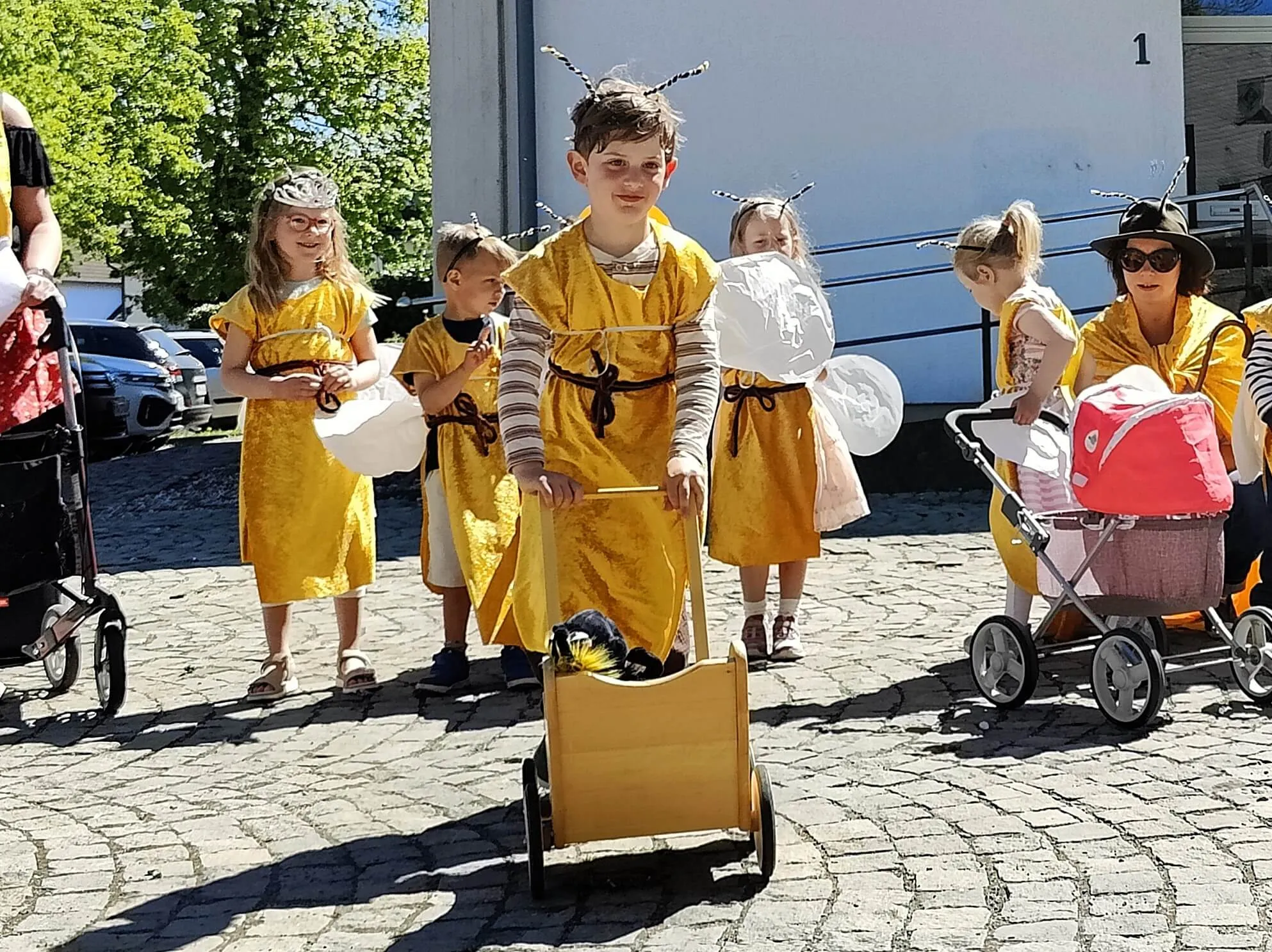Kinder in Bienenkostümen ziehen eine kleine Holzkutsche über einen Platz, umgeben von weiteren Personen und Kinderwagen.