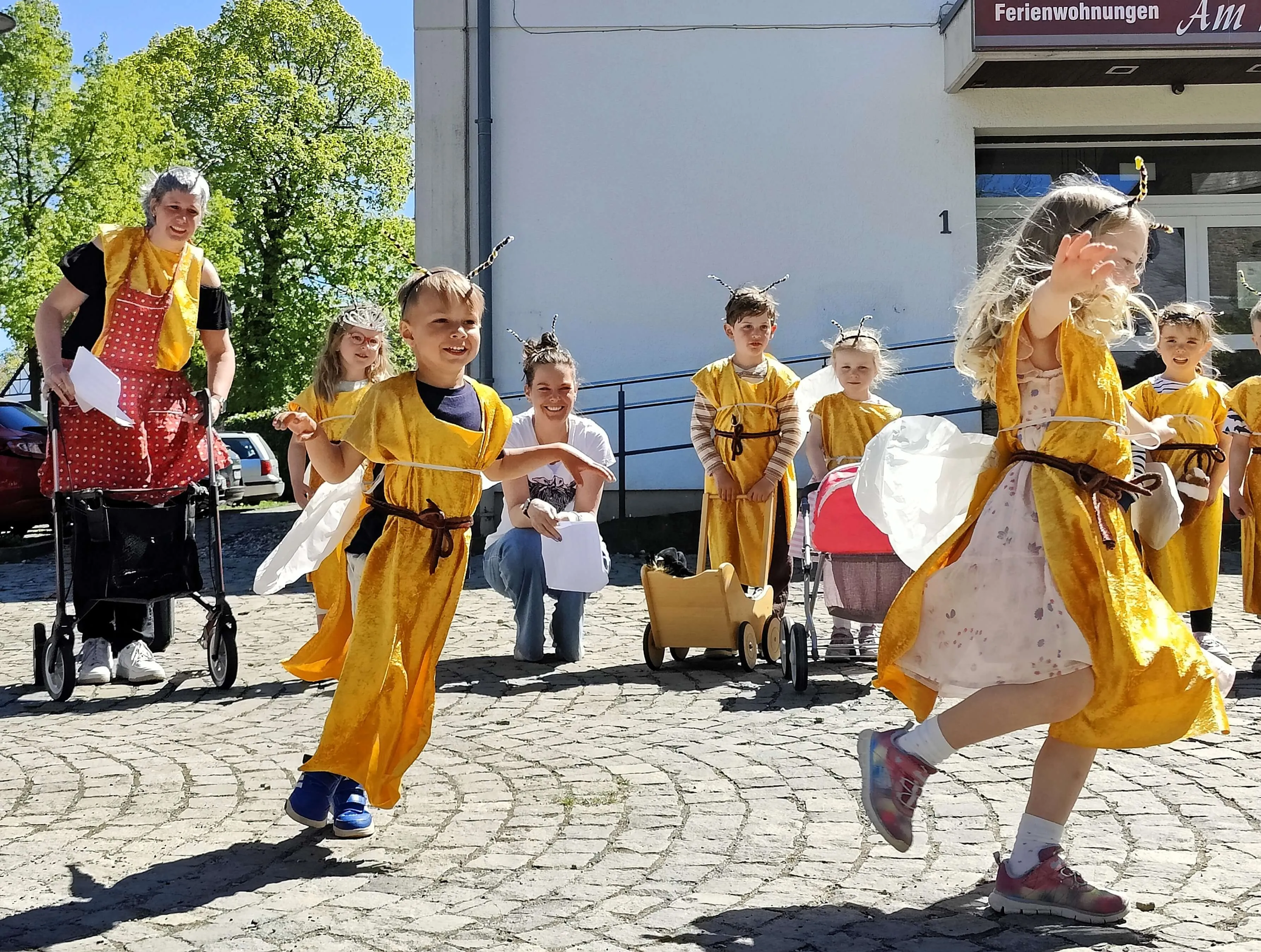 Kinder in gelben Kostümen mit Bienenflügeln tanzen und laufen auf einem gepflasterten Platz, während eine Frau im Rollstuhl und andere Zuschauer zusehen.