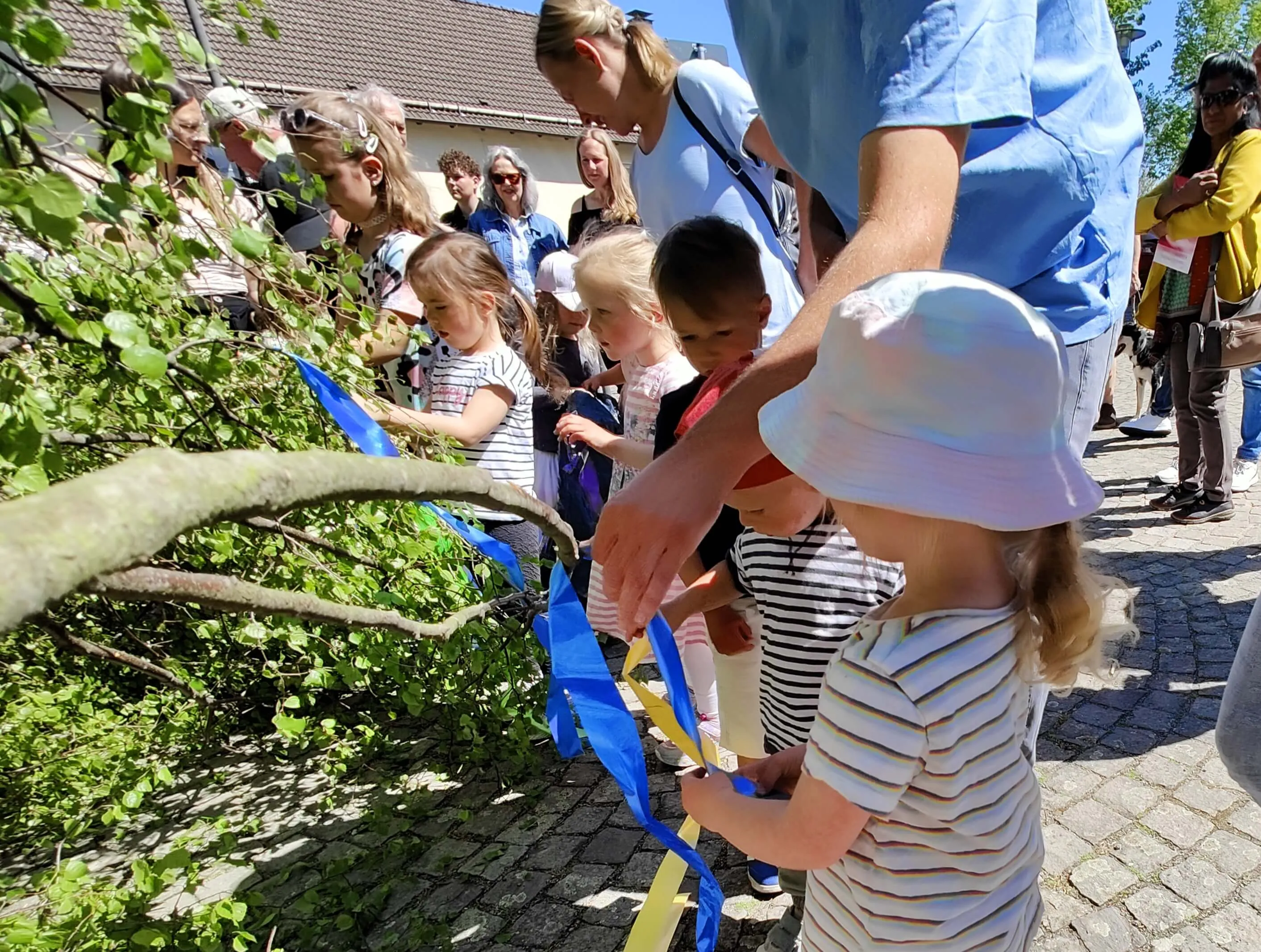 Kinder schneiden ein blaues Band mit einer Schere durch, umgeben von Erwachsenen und Zuschauern auf einem sonnigen Dorfplatz.