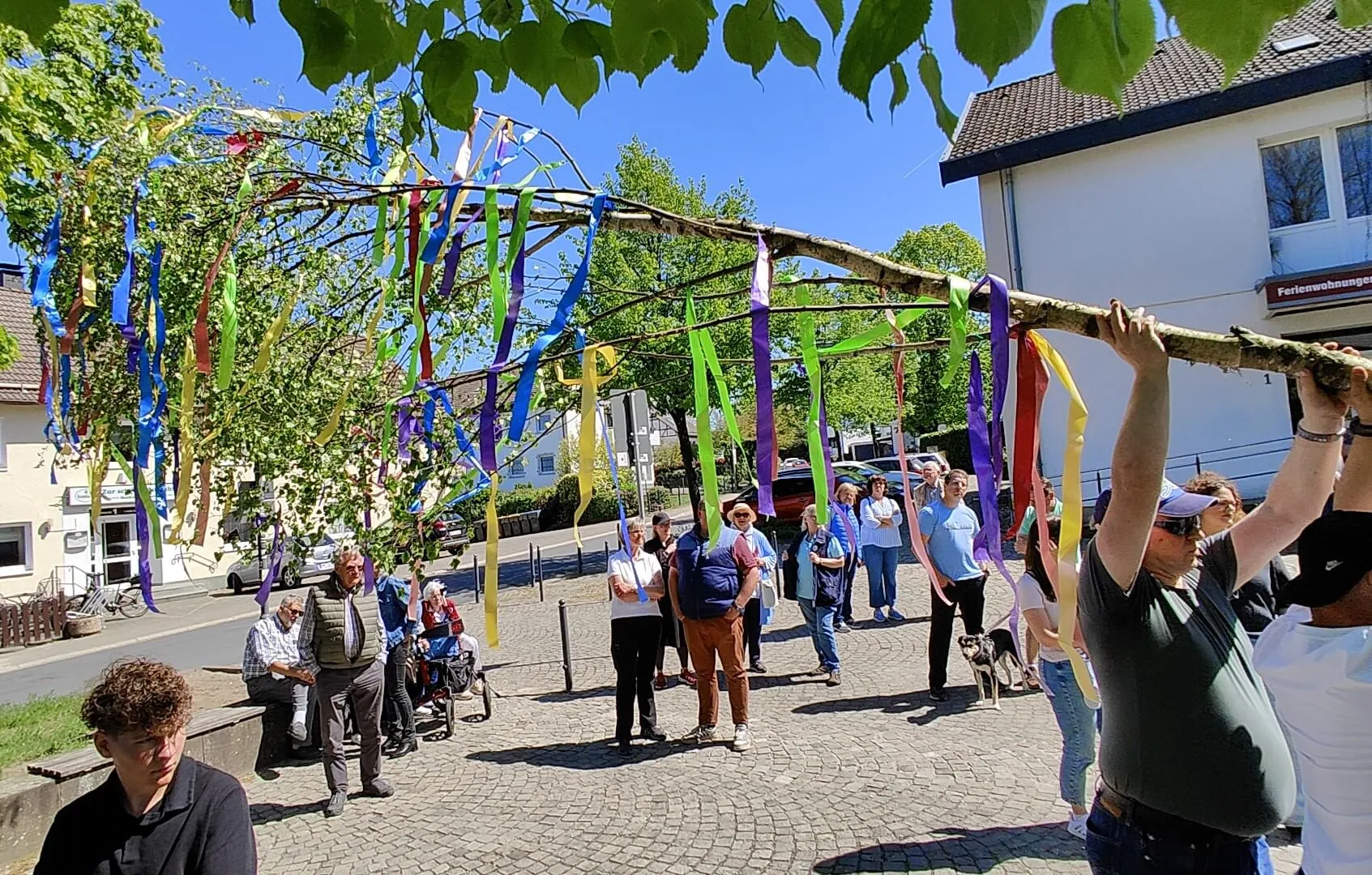 Eine bunt geschmückte Maistange wird von mehreren Personen auf einem Dorfplatz aufgestellt, umgeben von Zuschauern.