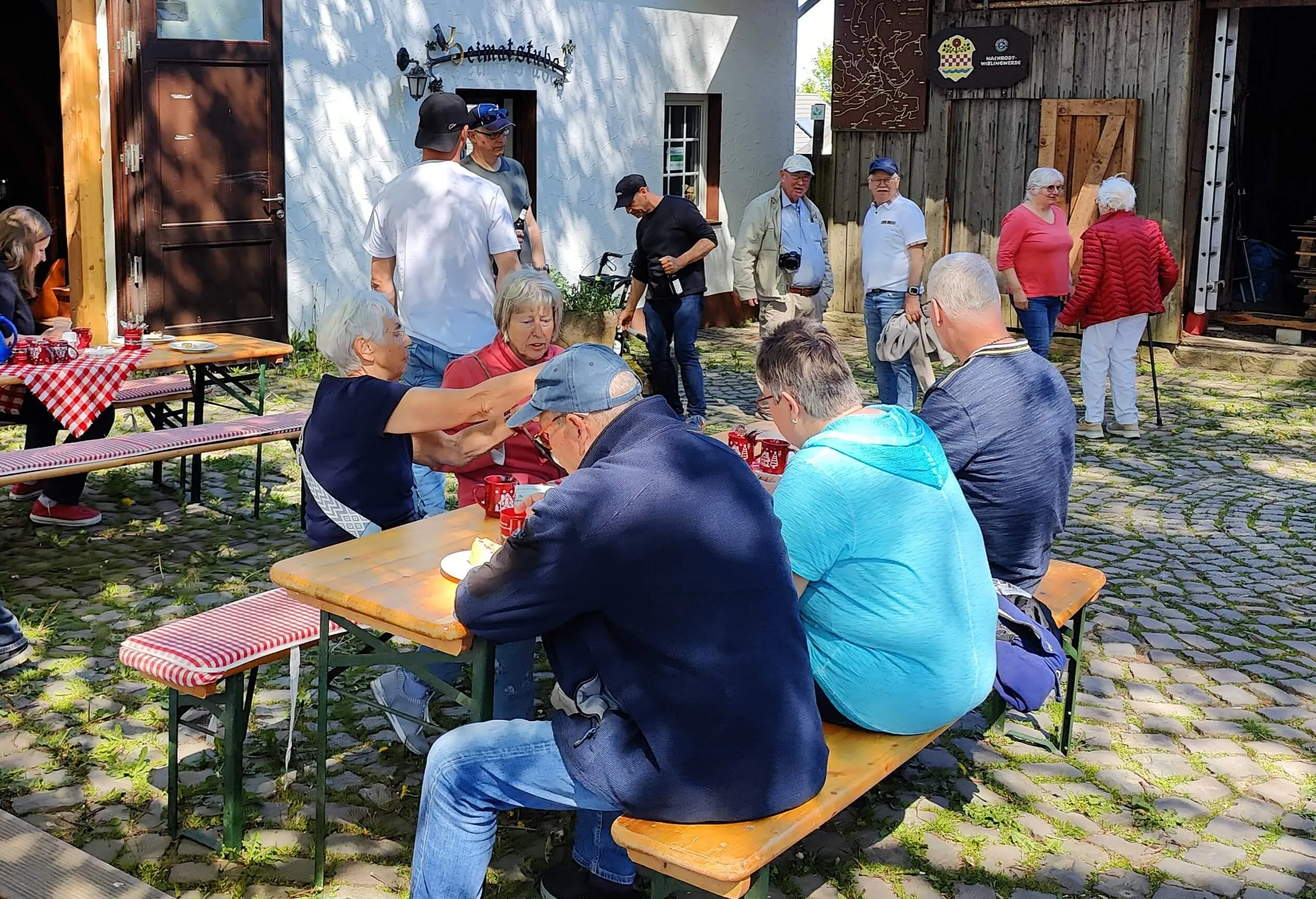 Menschen sitzen an Biertischgarnituren und essen im Freien, während andere im Hintergrund stehen und sich unterhalten. Im Hintergrund sind Gebäude und ein gepflasterter Platz zu sehen.