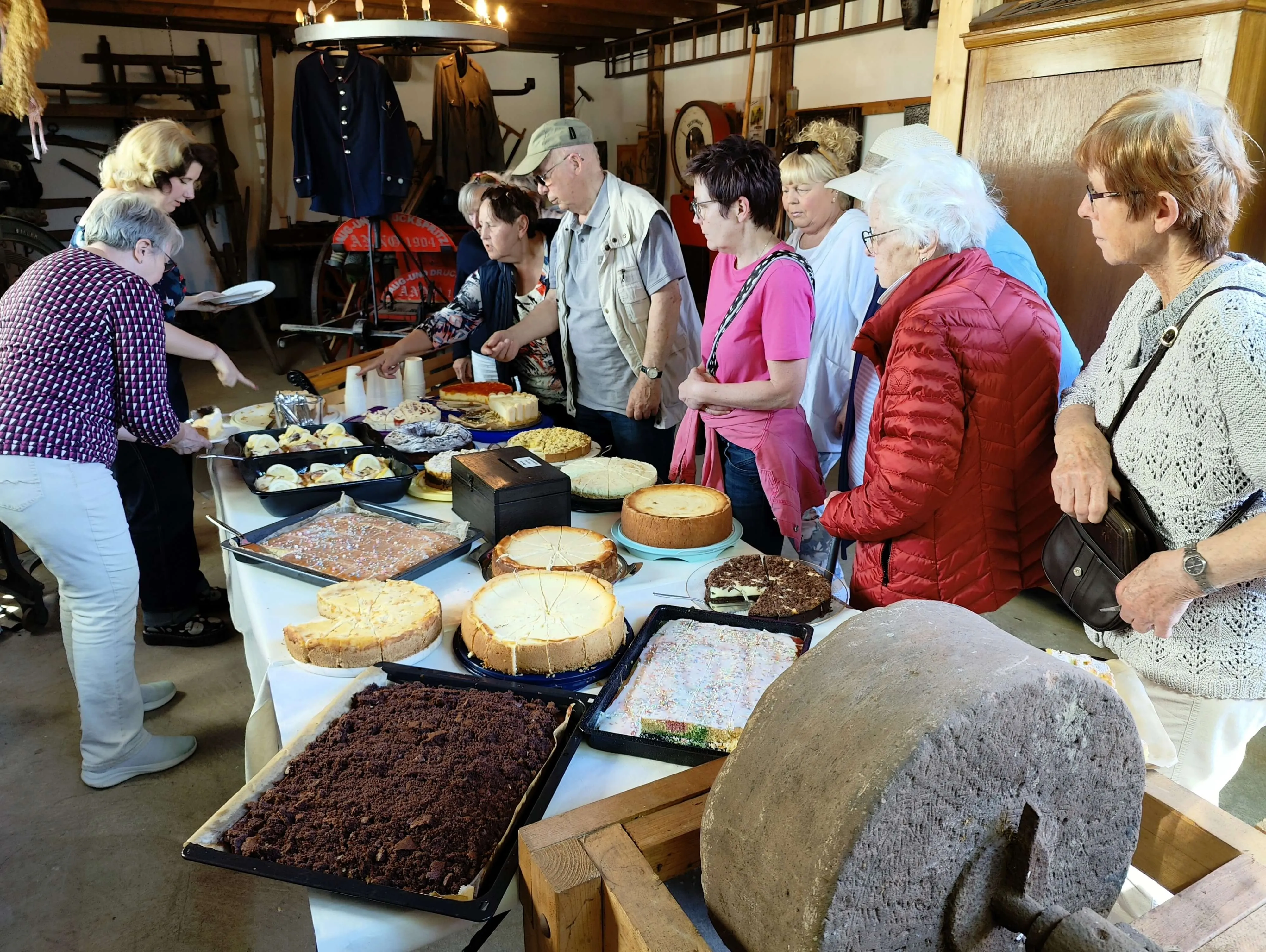 Eine Gruppe von Menschen steht um einen Tisch mit verschiedenen Kuchen und Torten auf einem Dorfplatz. Im Hintergrund sind historische Geräte und ein Gebäude zu sehen.