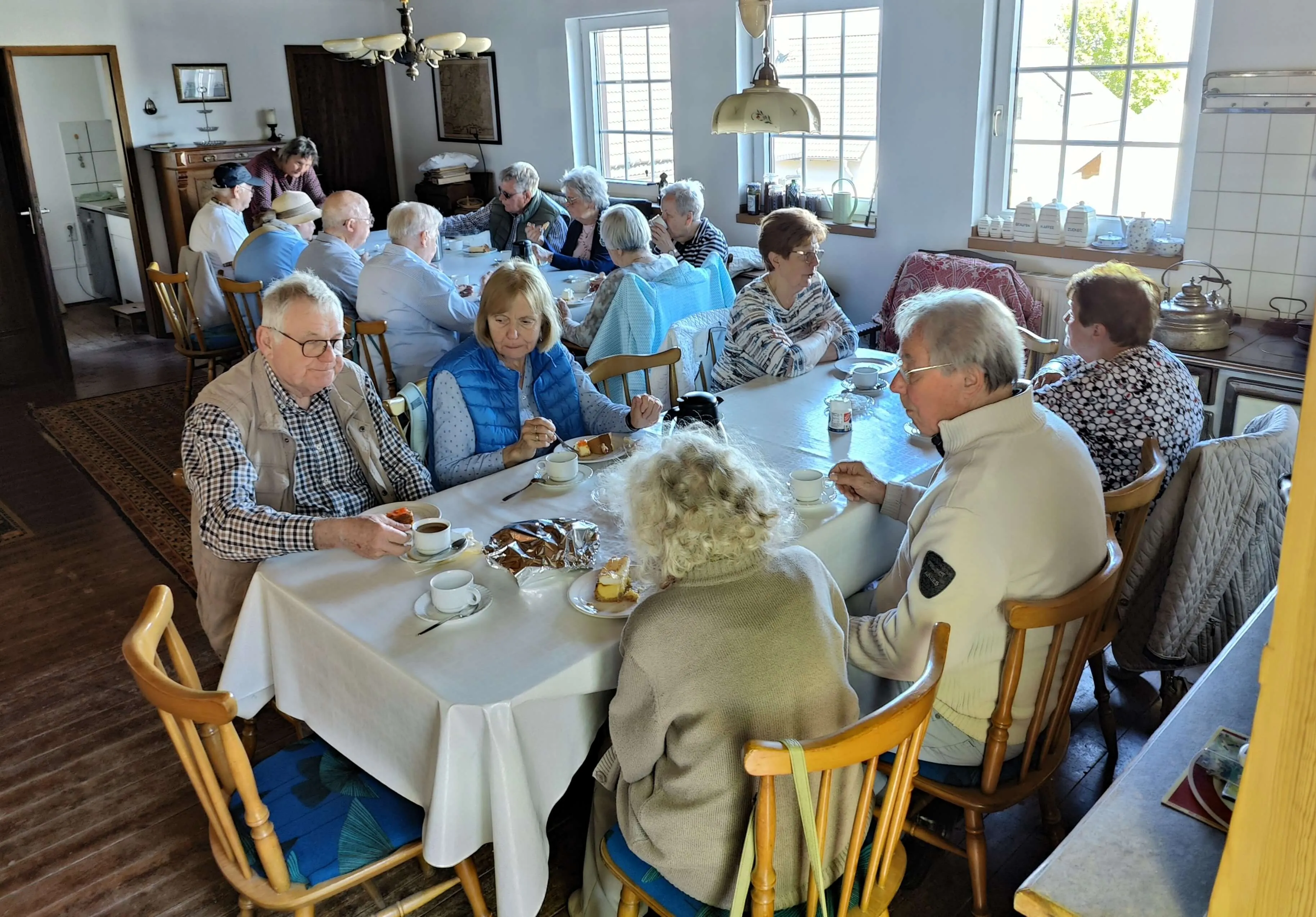 Eine Gruppe älterer Menschen sitzt an einem mit einer weißen Tischdecke bedeckten Tisch und isst Kuchen und trinkt Kaffee. Im Hintergrund weitere Personen und ein helles Rauminnere.
