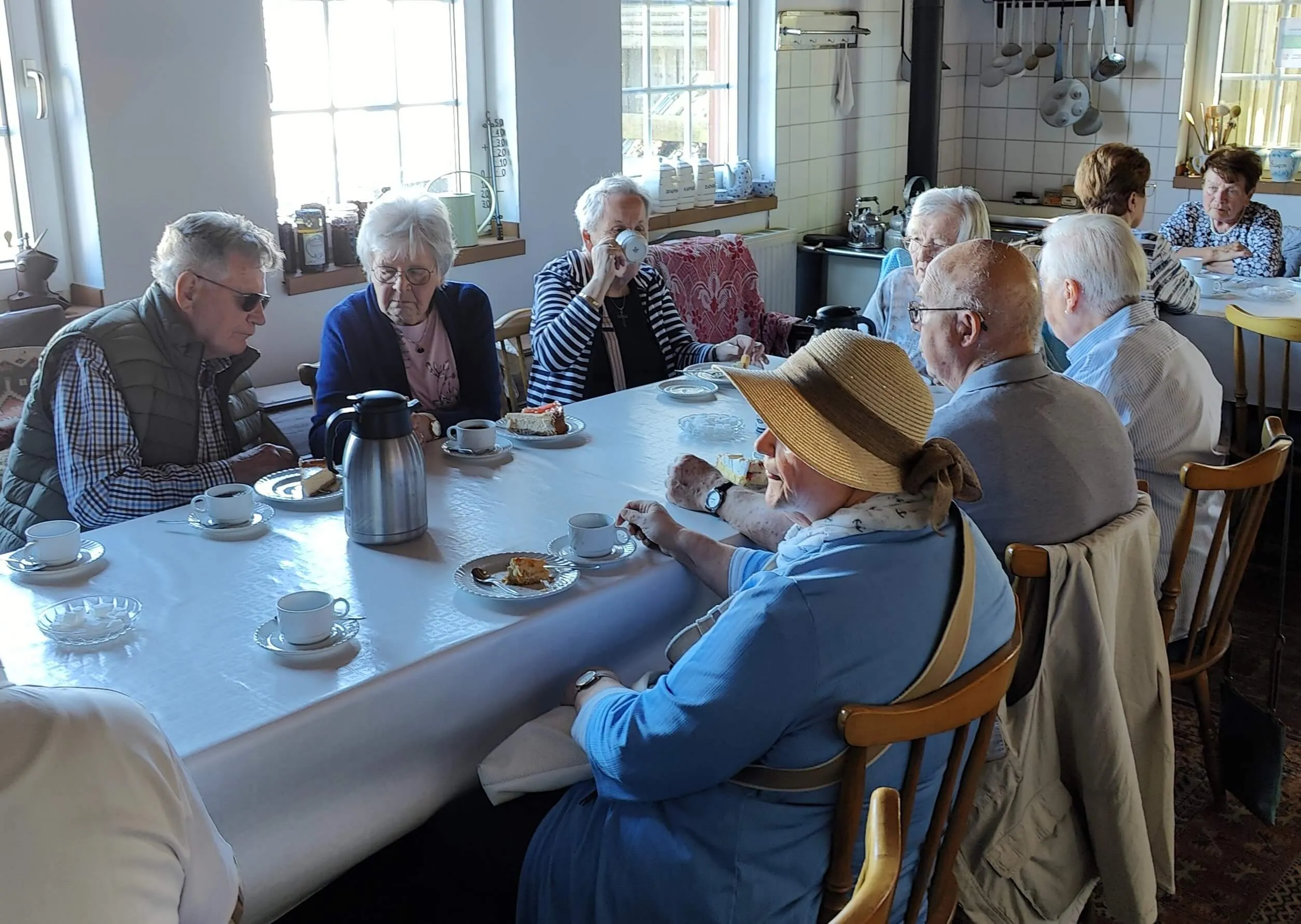 Eine Gruppe älterer Menschen sitzt an einem langen Tisch und genießt Kaffee und Kuchen. Im Hintergrund sind weitere Personen und eine Küche zu sehen.