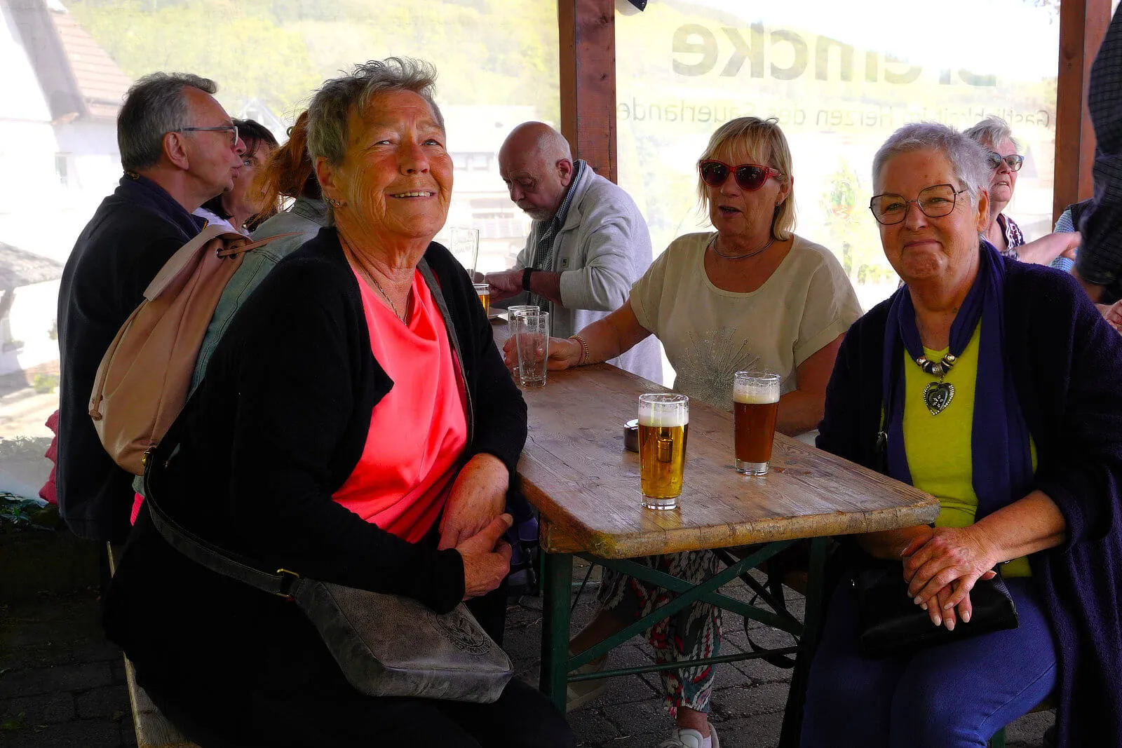 Mehrere Menschen sitzen an einem Holztisch im Freien, trinken Bier und unterhalten sich. Im Hintergrund sind weitere Personen und ein Gebäude zu sehen.