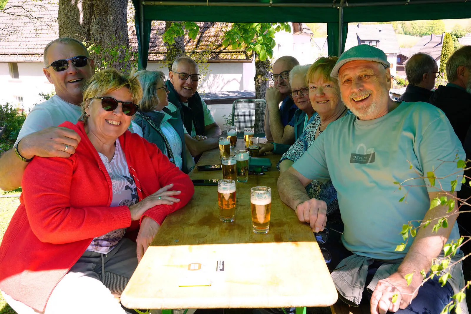 Eine Gruppe von Menschen sitzt an einem Holztisch unter einem schattigen Pavillon und genießt Bier. Sie lächeln und unterhalten sich bei einem Frühlingsfest im Freien.