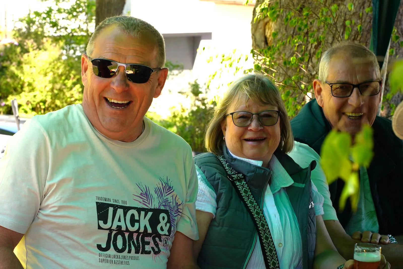 Drei lächelnde Menschen unterschiedlichen Alters sitzen im Freien zusammen. Ein Mann mit Sonnenbrille trägt ein T-Shirt der Marke Jack & Jones, während eine Frau mit Brille und ein älterer Mann mit Glas in der Hand neben ihr sitzen.