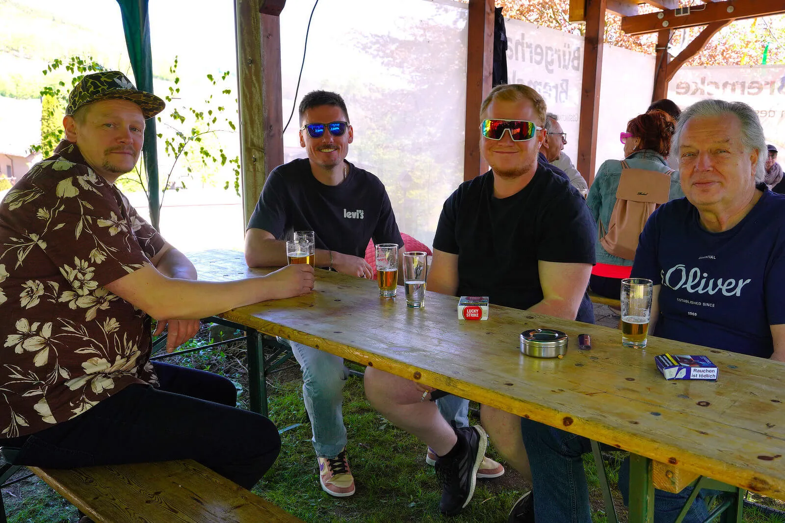 Vier Männer sitzen an einem Holztisch im Freien, Biergläser und eine Schachtel Zigaretten stehen darauf. Im Hintergrund sind weitere Personen und ein grüner Garten zu sehen.