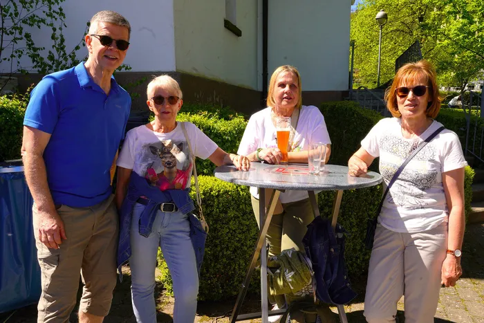 Vier Menschen stehen und sitzen im Freien um einen runden Tisch, Getränke dabei. Im Hintergrund ein Gebäude und Bäume.
