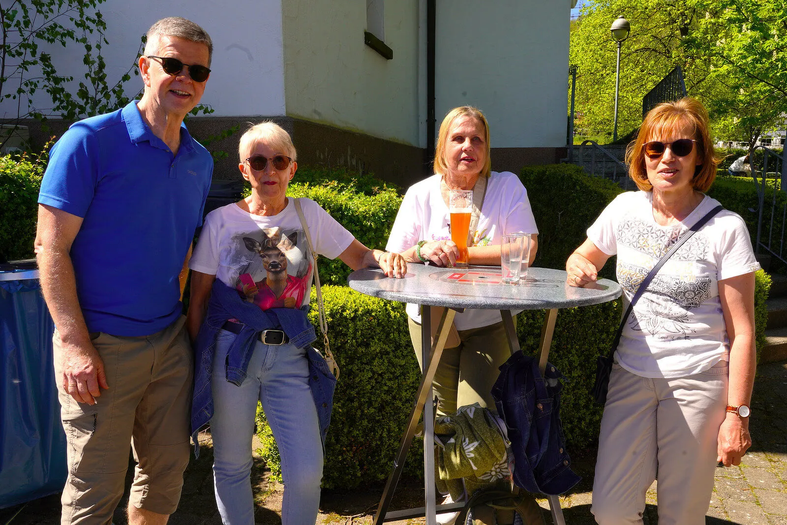 Vier Menschen stehen und sitzen im Freien um einen runden Tisch, Getränke dabei. Im Hintergrund ein Gebäude und Bäume.