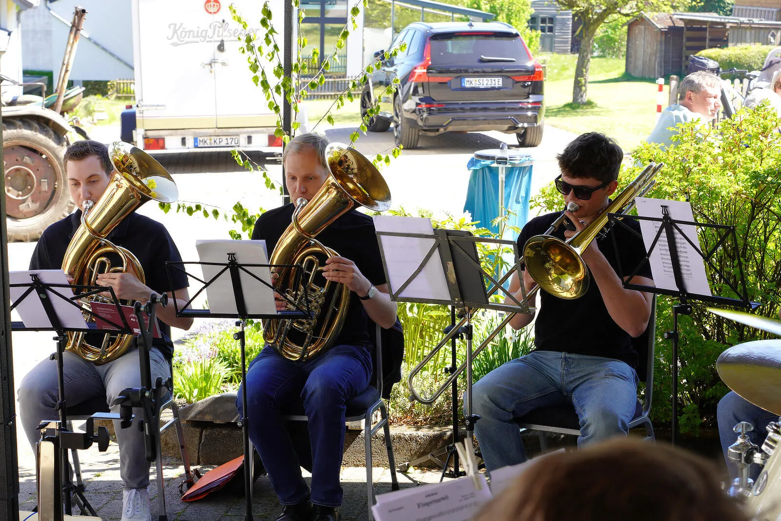 Vier Bläser spielen auf Tuba, Posaune und Horn im Freien vor Notenständern. Im Hintergrund ein parkender grauer Wagen und ein Anhänger.