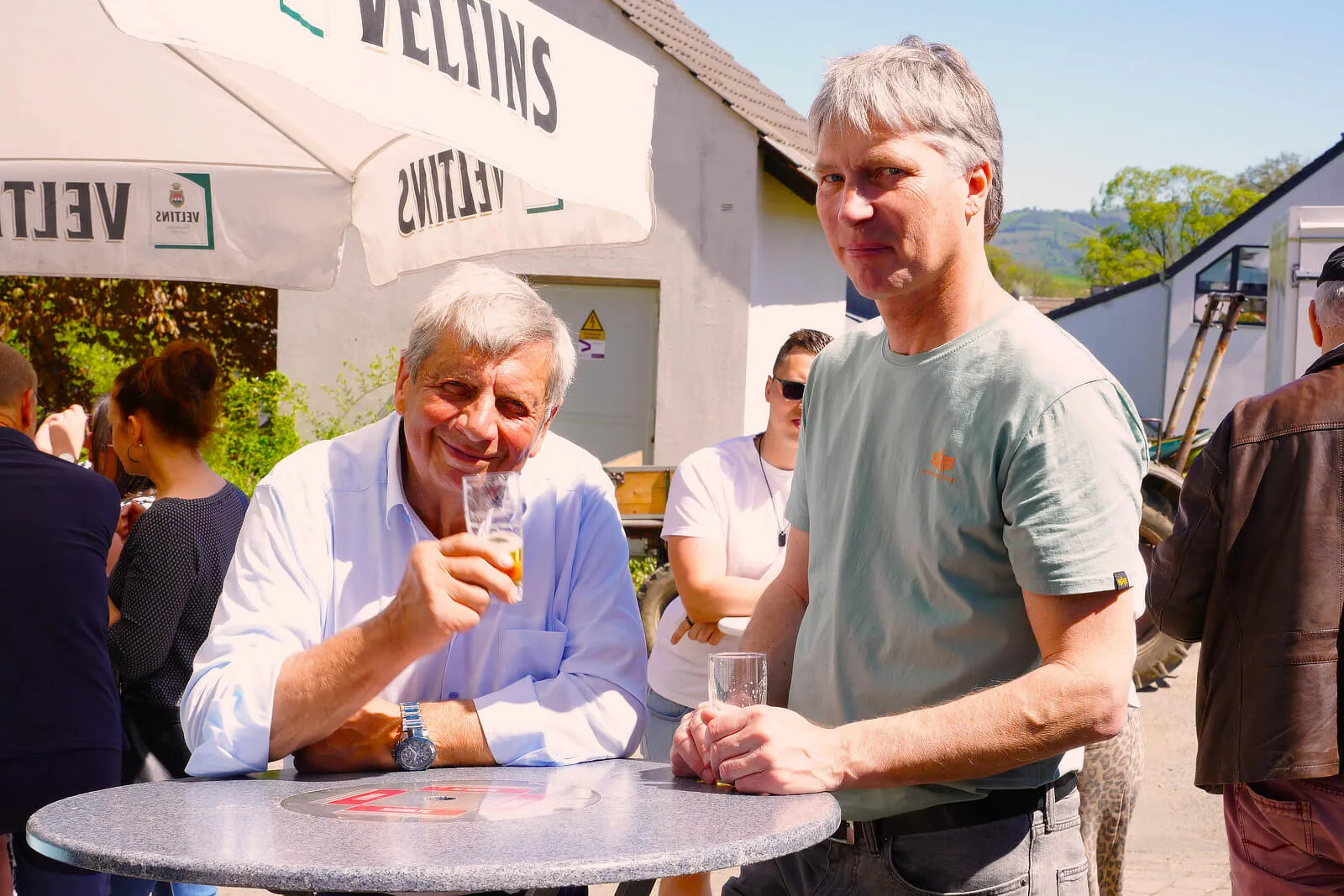 Zwei Männer unterschiedlichen Alters stehen an einem Tisch im Freien und halten Biergläser. Im Hintergrund sind weitere Personen und ein Gebäude mit einem Sonnenschirm zu sehen.