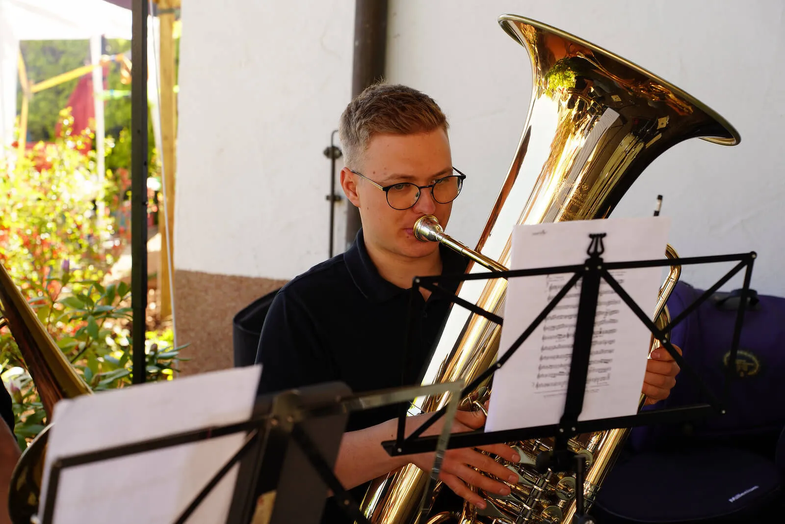 Ein junger Mann mit Brille spielt auf einer goldenen Tuba, während er Notenblätter vor sich hat. Im Hintergrund sind blühende Pflanzen zu sehen.