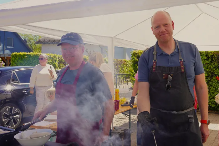 Zwei Männer stehen unter einem weißen Zelt und grillen. Im Hintergrund stehen weitere Personen und ein Auto.
